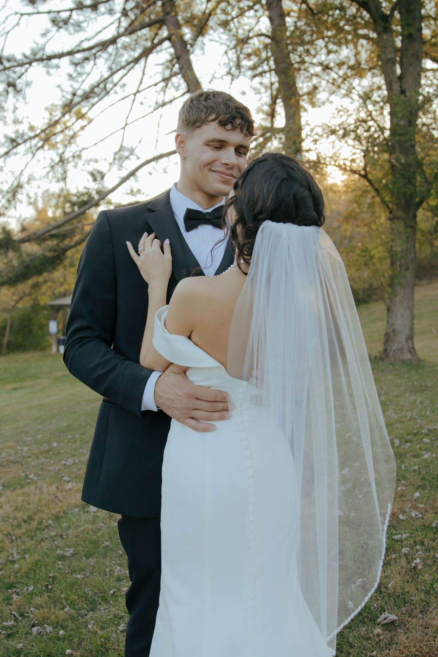 Smiling groom in black tuxedo embraces bride in white wedding gown among autumn trees at sunset.