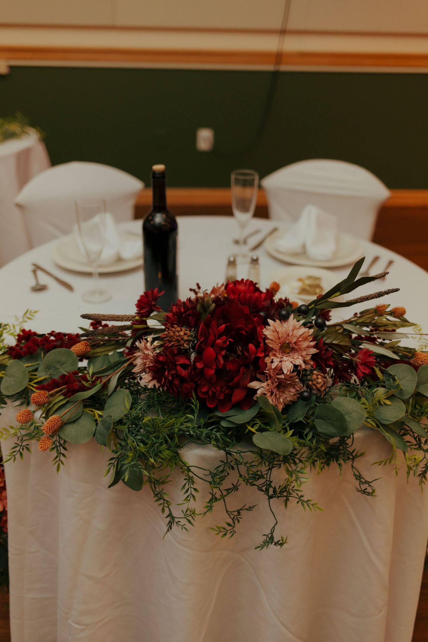 Sweetheart table centerpiece with deep red dahlias, greenery, and wine bottle.