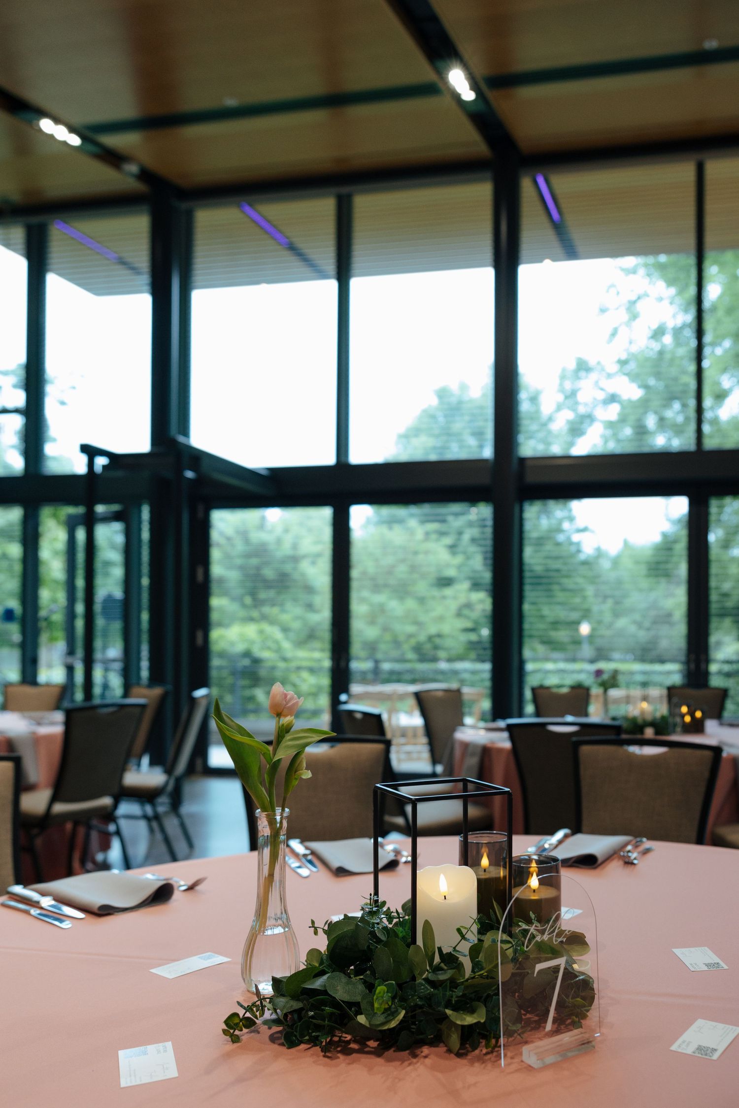 Elegant wedding reception centerpiece with white candles, greenery, and a tulip in a glass vase, set in a modern venue with large windows.