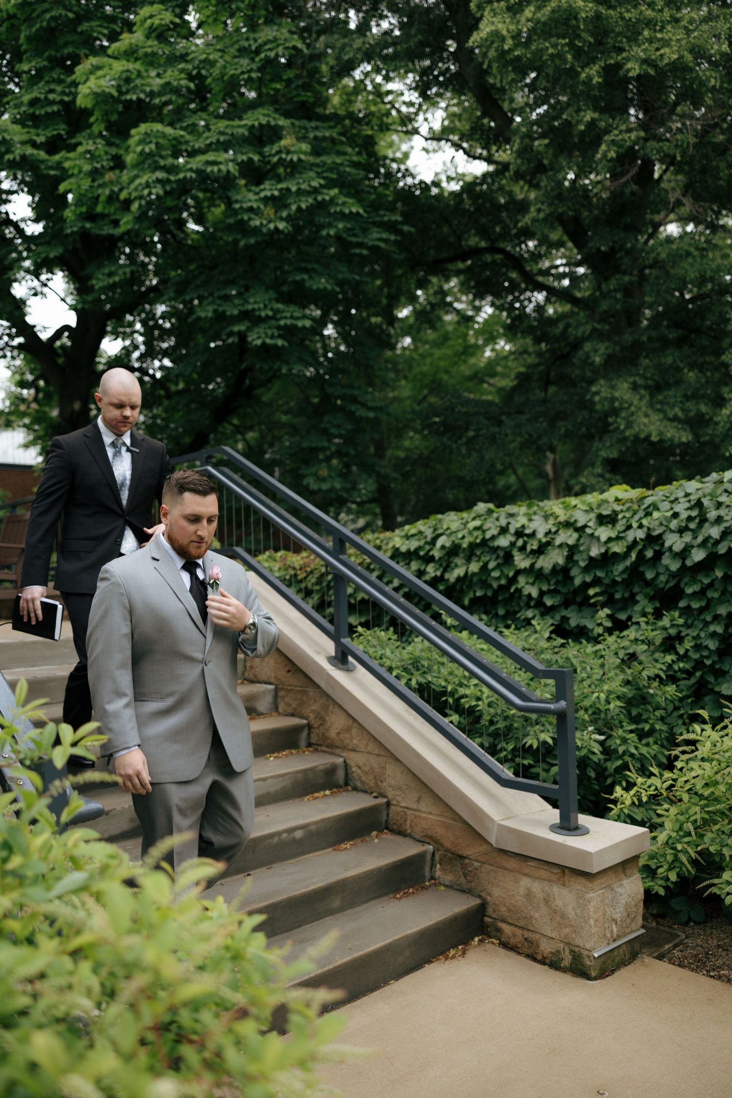 Two men in suits descend outdoor stone steps surrounded by lush green trees and hedges at a wedding venue.