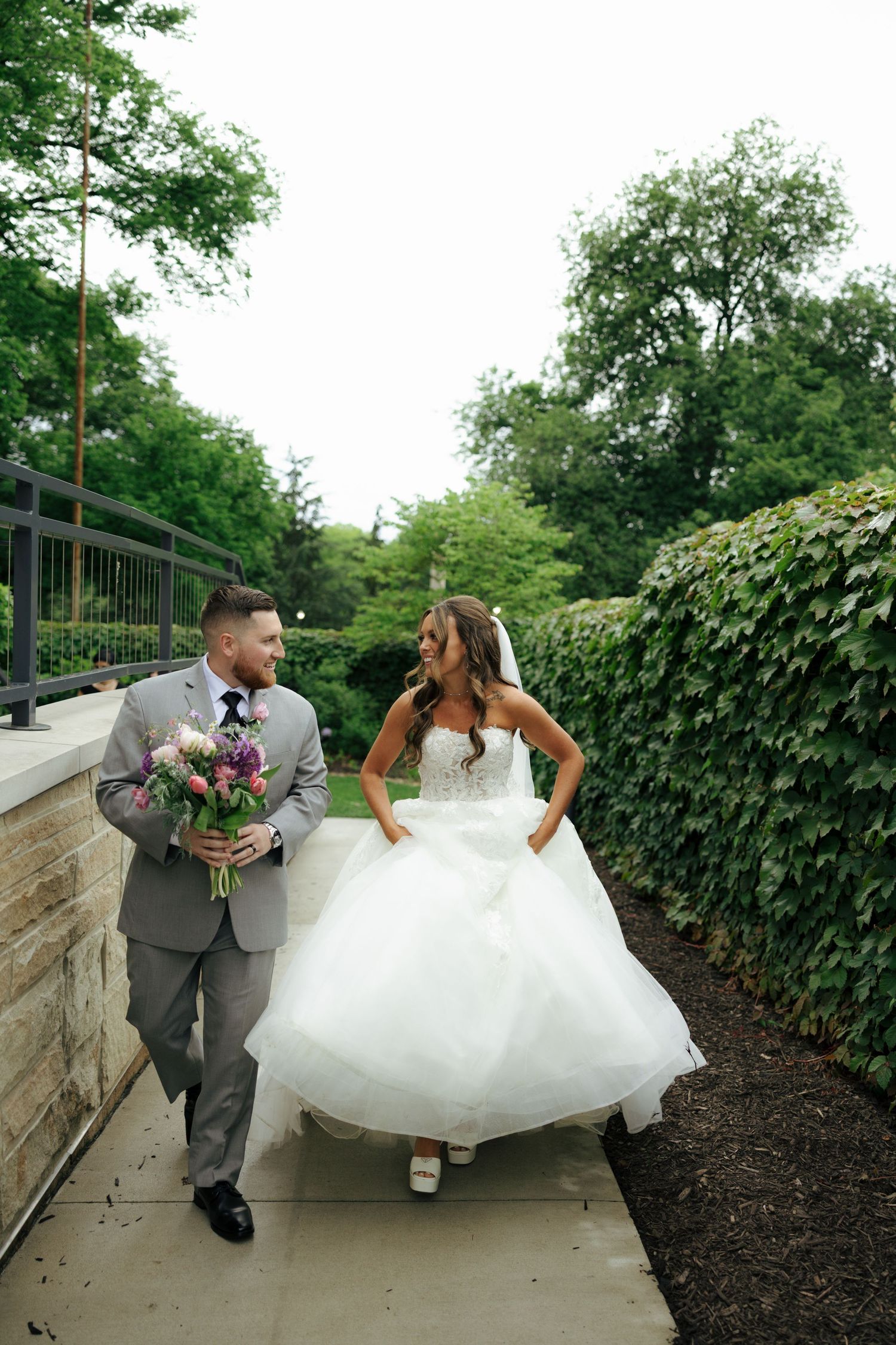 Bride and groom walking together outdoors, smiling at each other, groom holding purple bouquet, lush green garden backdrop.