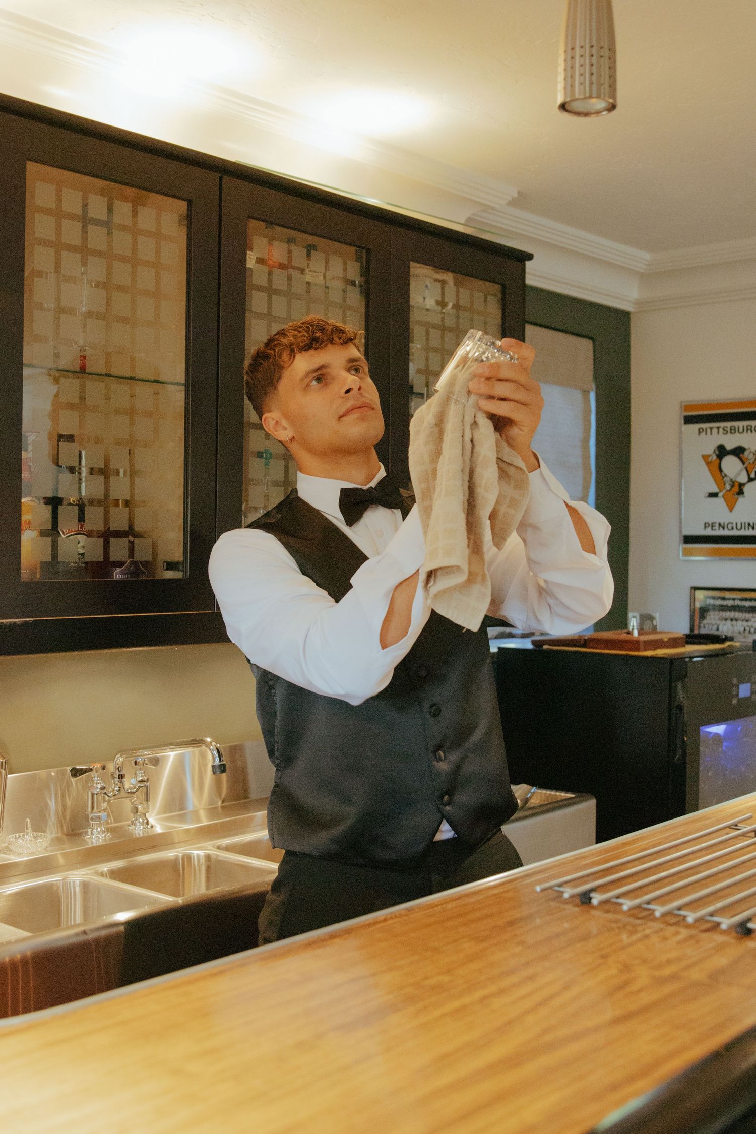Young man in vest and bow tie polishing a glass behind a wooden bar counter.