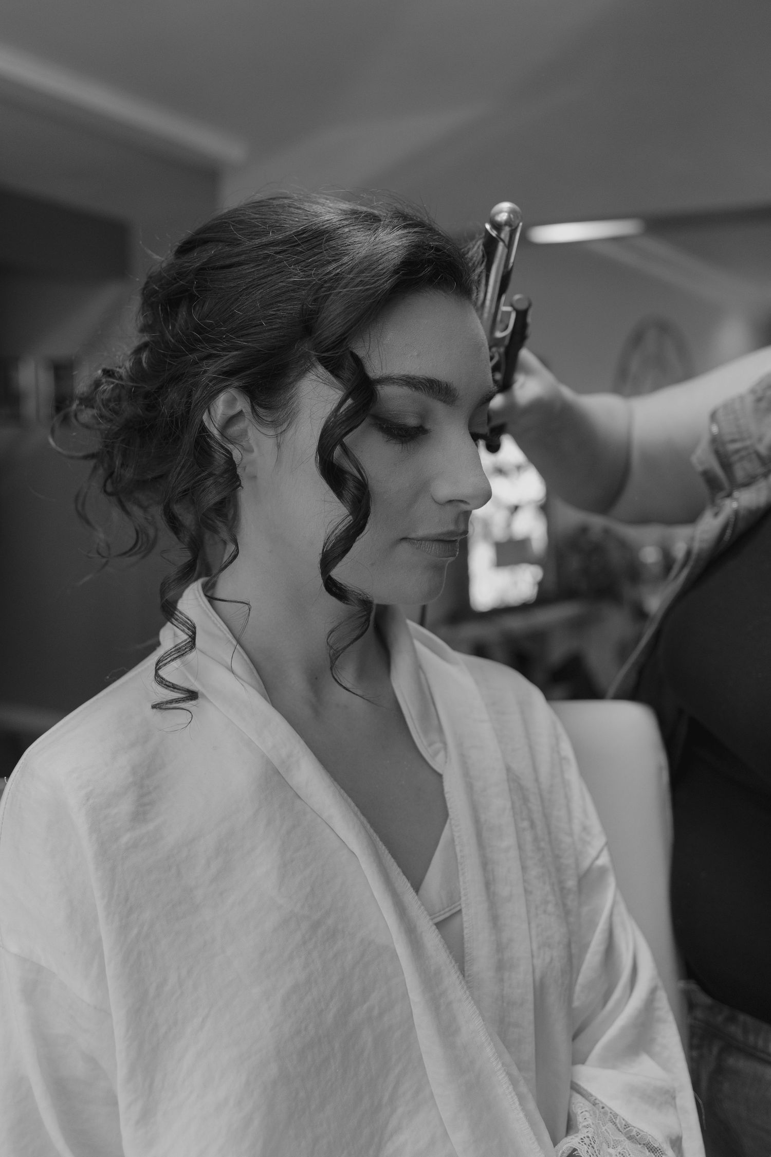 Black and white photo of woman getting curly hair styled with curling iron before wedding ceremony.
