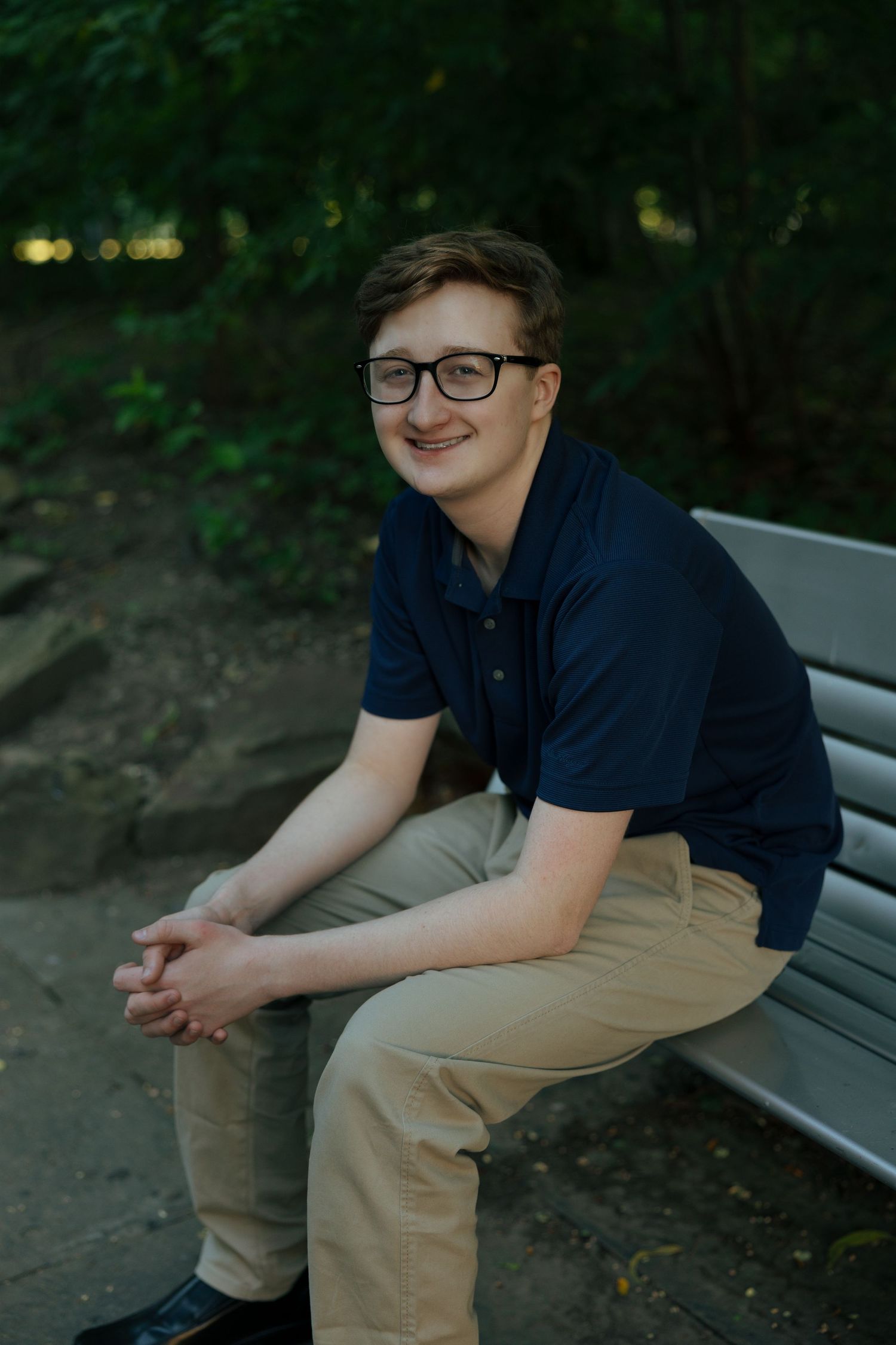 Teenage boy in navy polo and khakis sitting on a park bench, smiling with green trees in background.