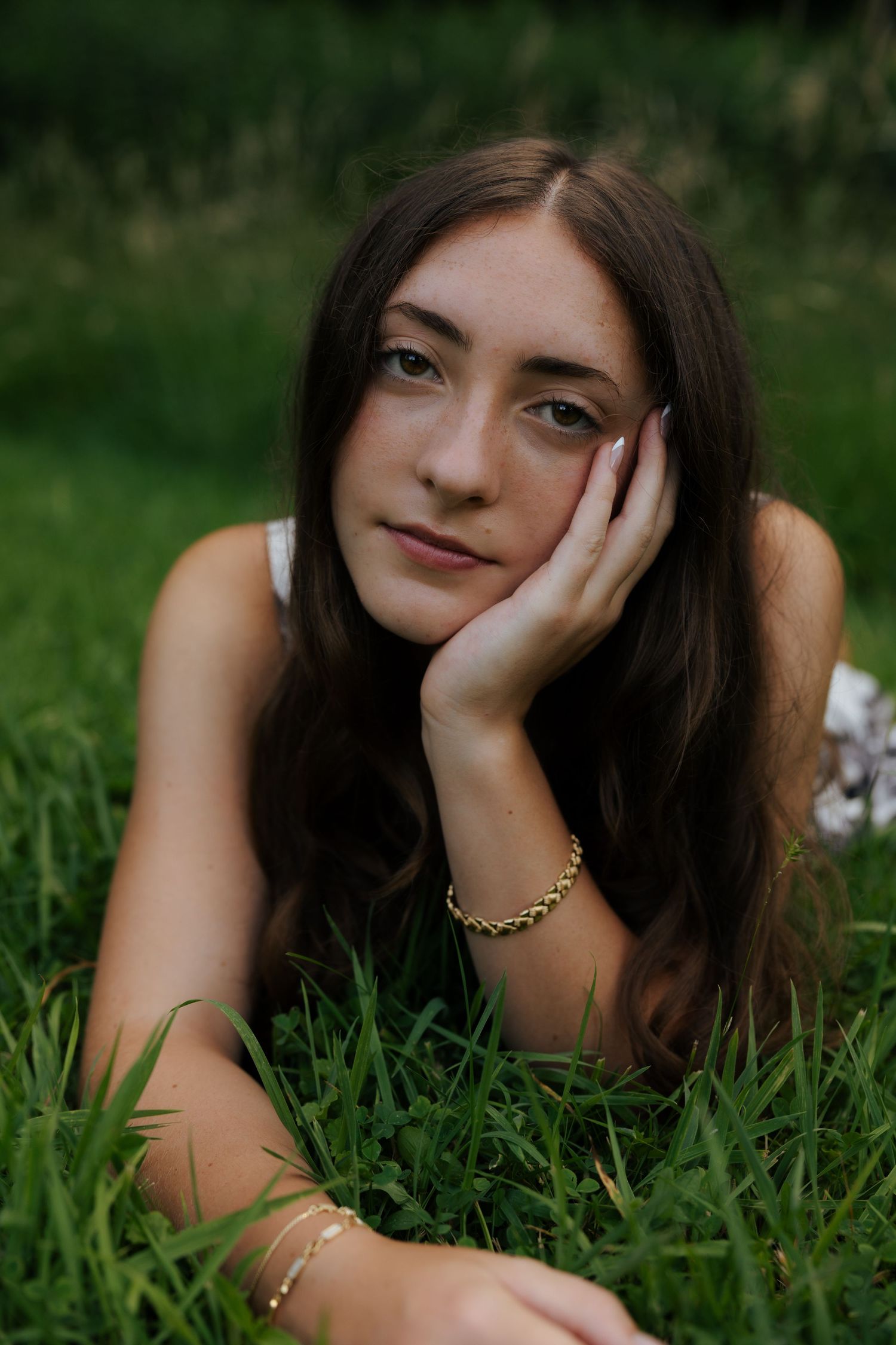 Young woman with dark hair lying in green grass, resting cheek on hand, wearing gold bracelets.