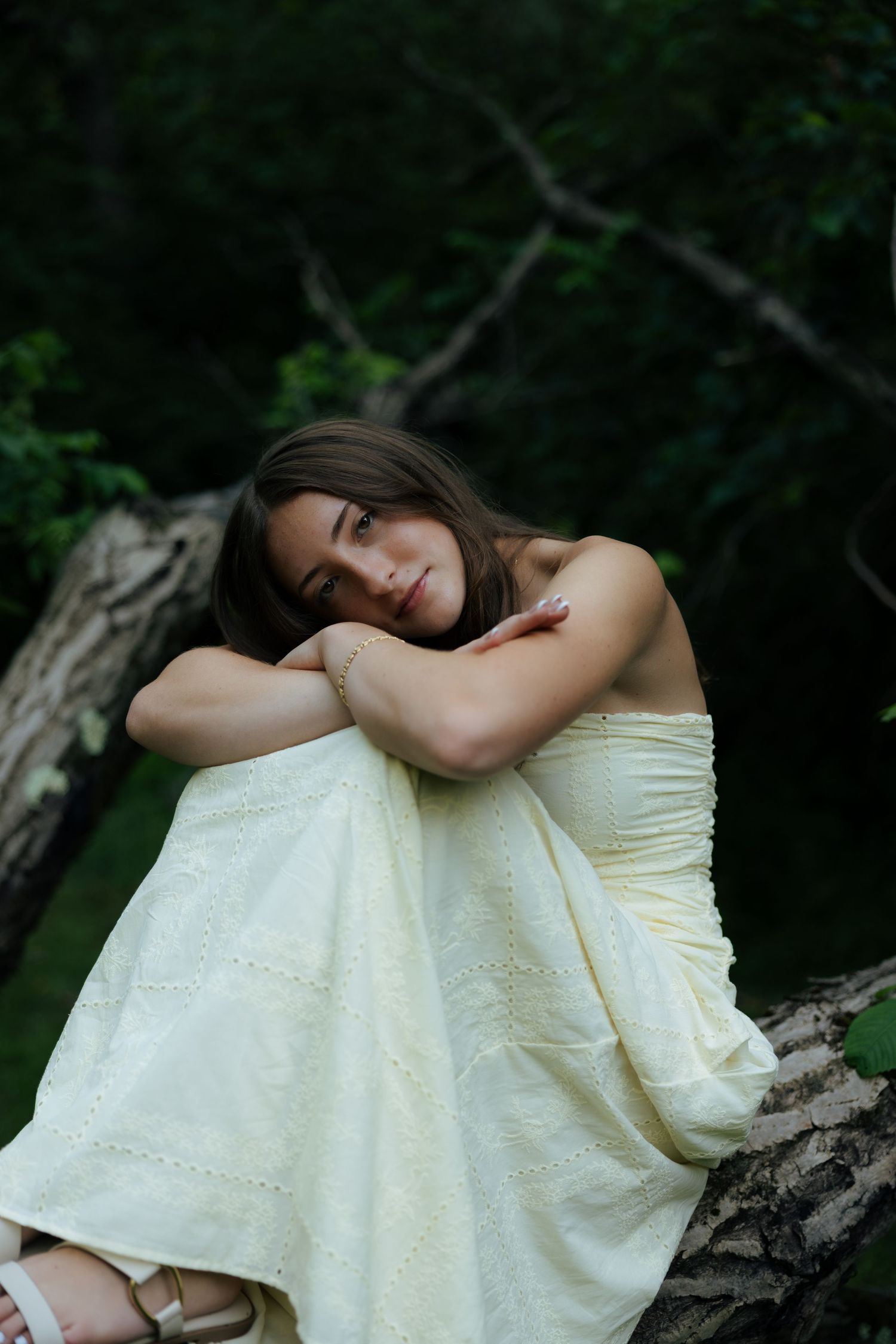 Young woman in yellow strapless dress resting on fallen tree in dark forest.