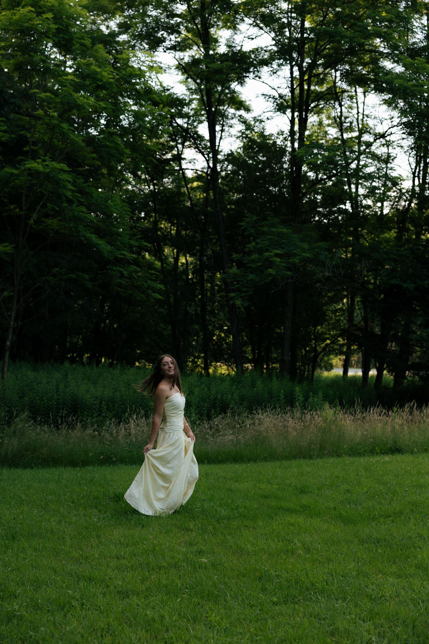 Young woman in flowing cream gown twirling in lush green meadow with tall trees in background.