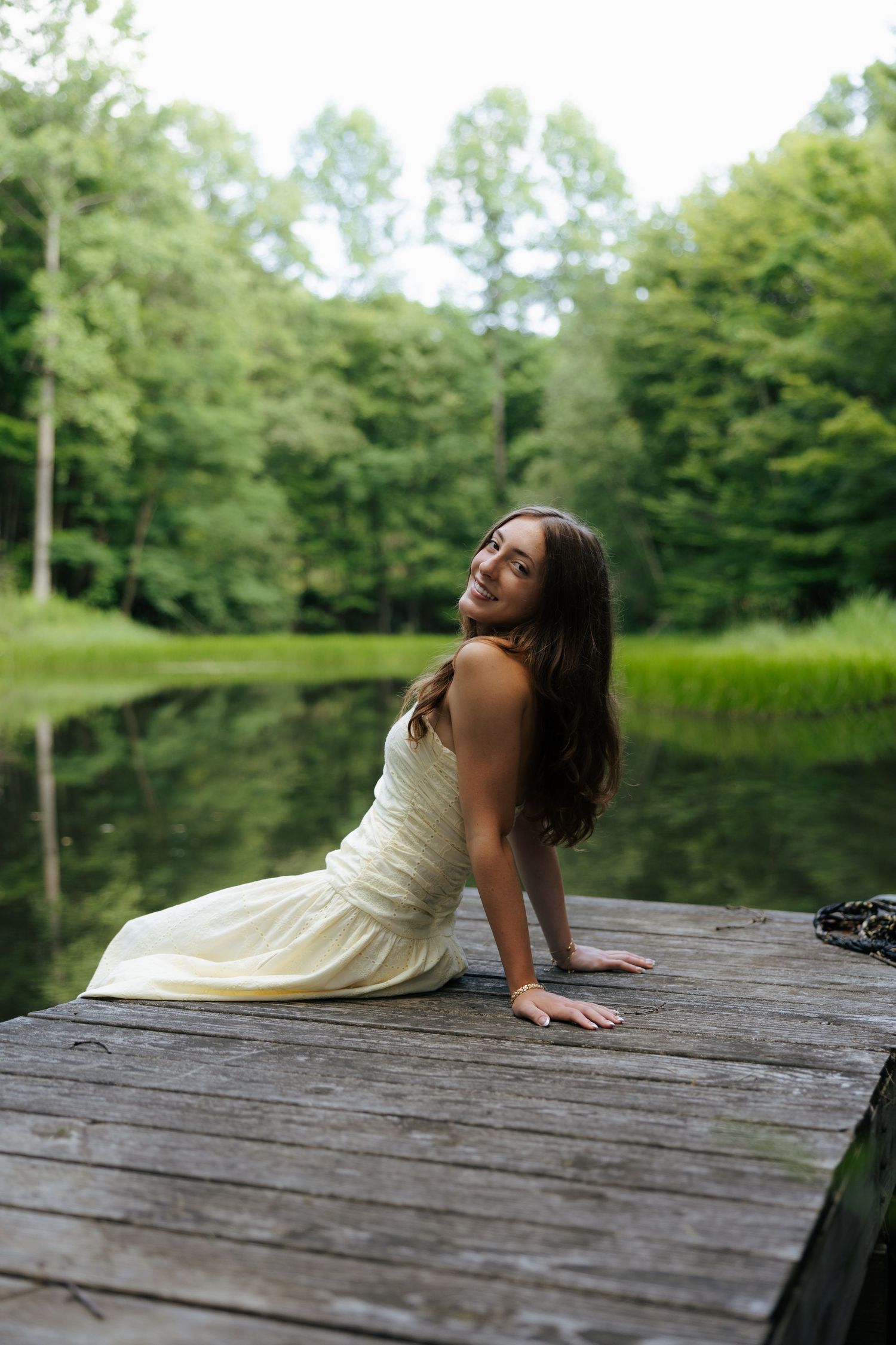 Young woman in white dress sitting gracefully on wooden dock with serene green lake backdrop.