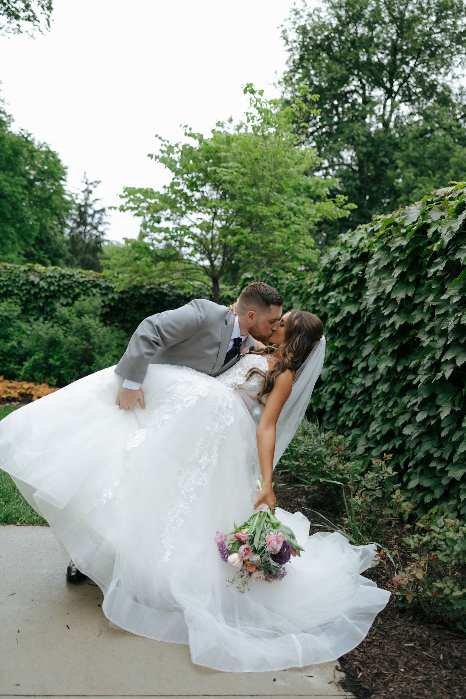 Groom dips bride in stunning white lace ballgown for romantic kiss in lush garden setting.