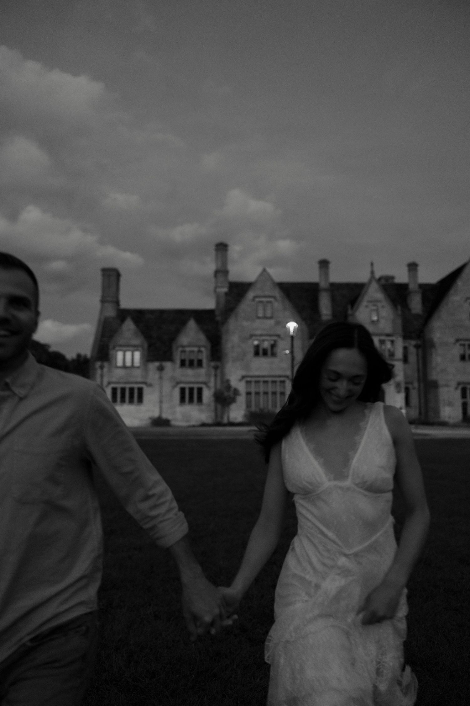 Black and white photo of couple holding hands walking before large stone manor at dusk.