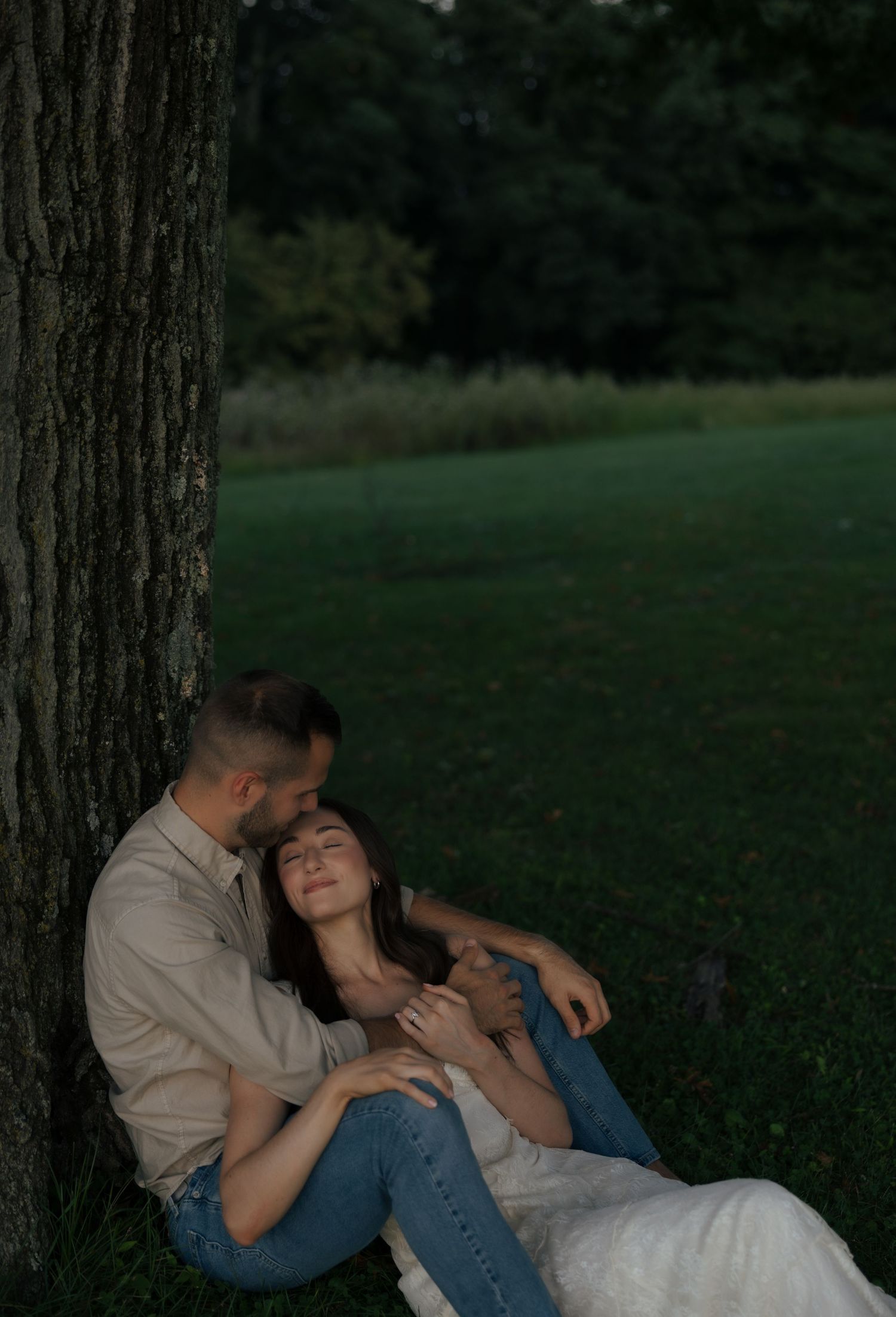 Engaged couple cuddles romantically against large tree trunk on lush green lawn at dusk.