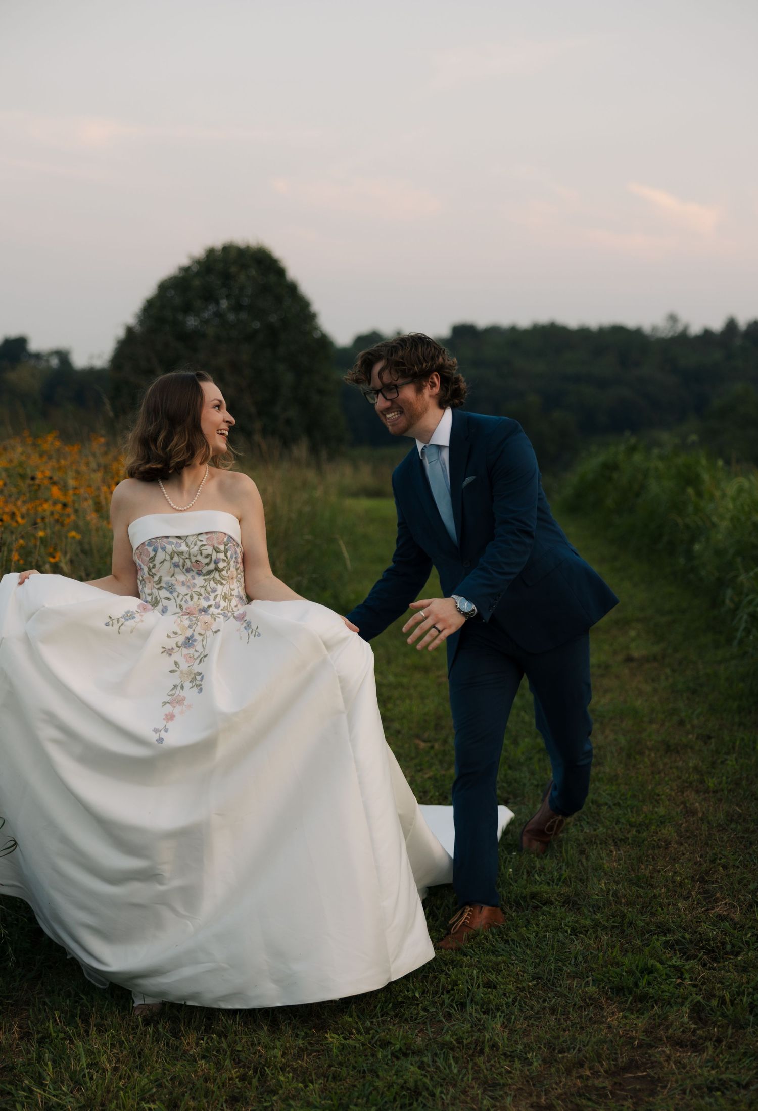 Laughing bride and groom holding hands while walking along a grassy path surrounded by wildflowers at sunset.