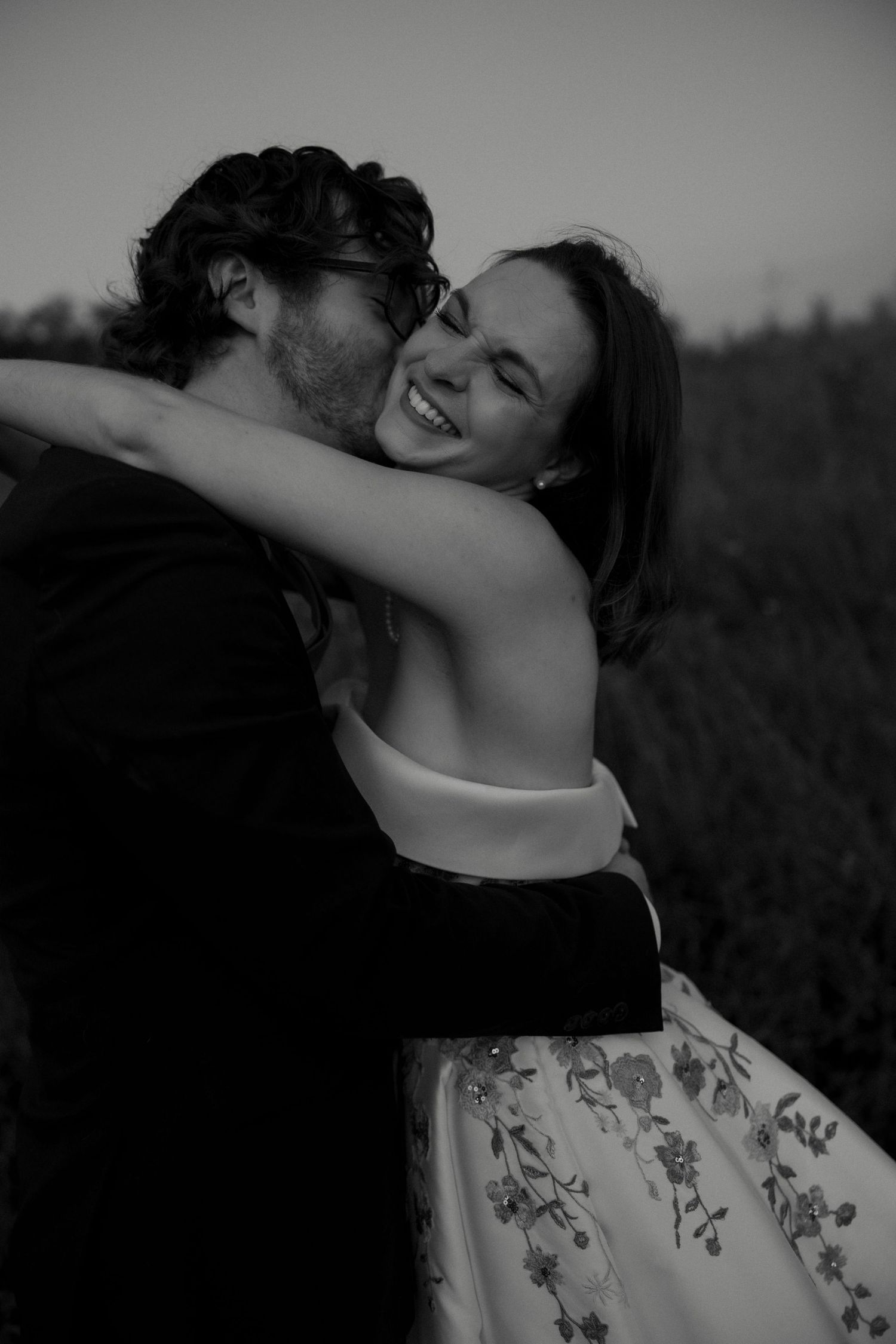 Black and white photo of laughing bride embracing groom outdoors during wedding portrait session.