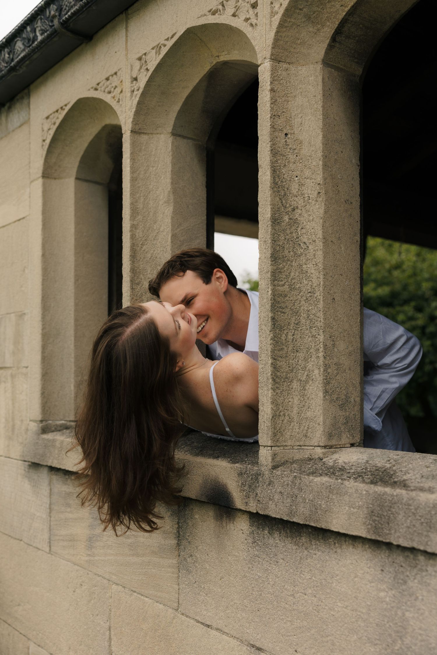 Laughing couple sharing a playful kiss through stone archway openings on a historic building.