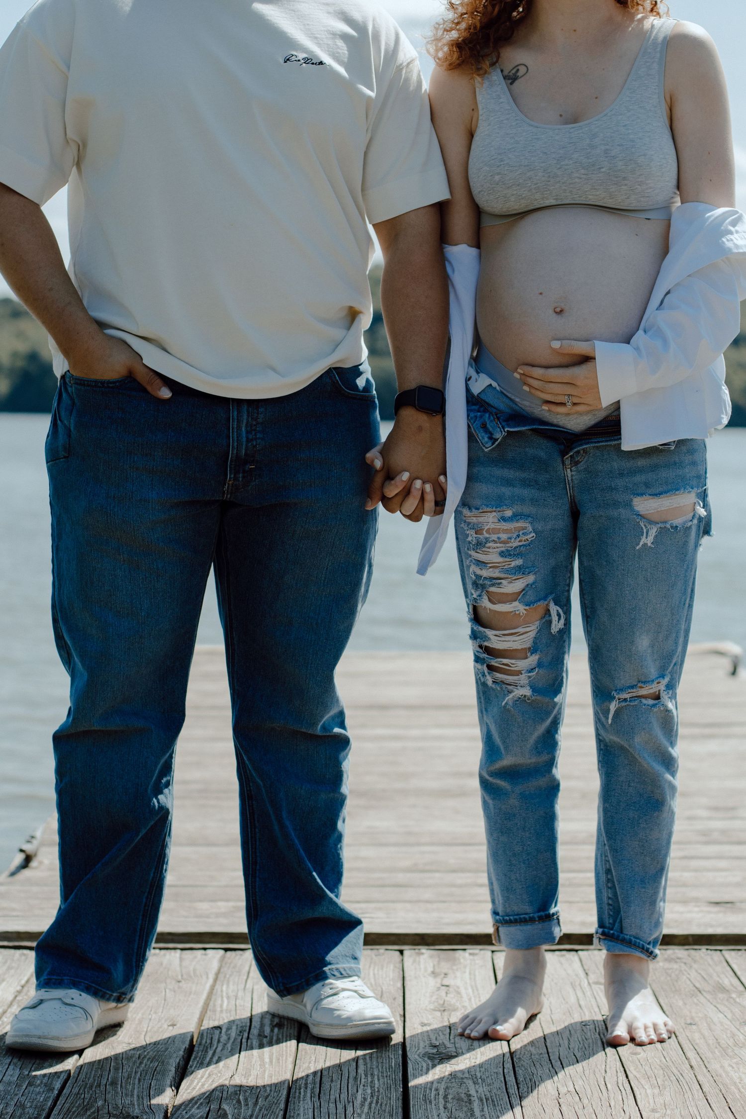 Pregnant couple holding hands on a wooden dock, woman in ripped jeans and crop top cradling baby bump.