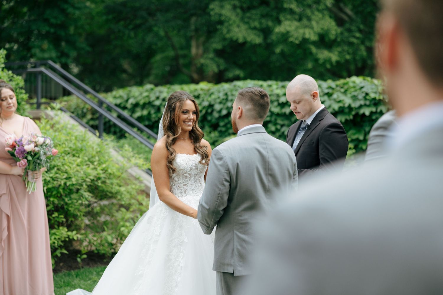 Bride and groom exchange vows outdoors during a wedding ceremony, with a officiant and bridesmaid in blush pink nearby.