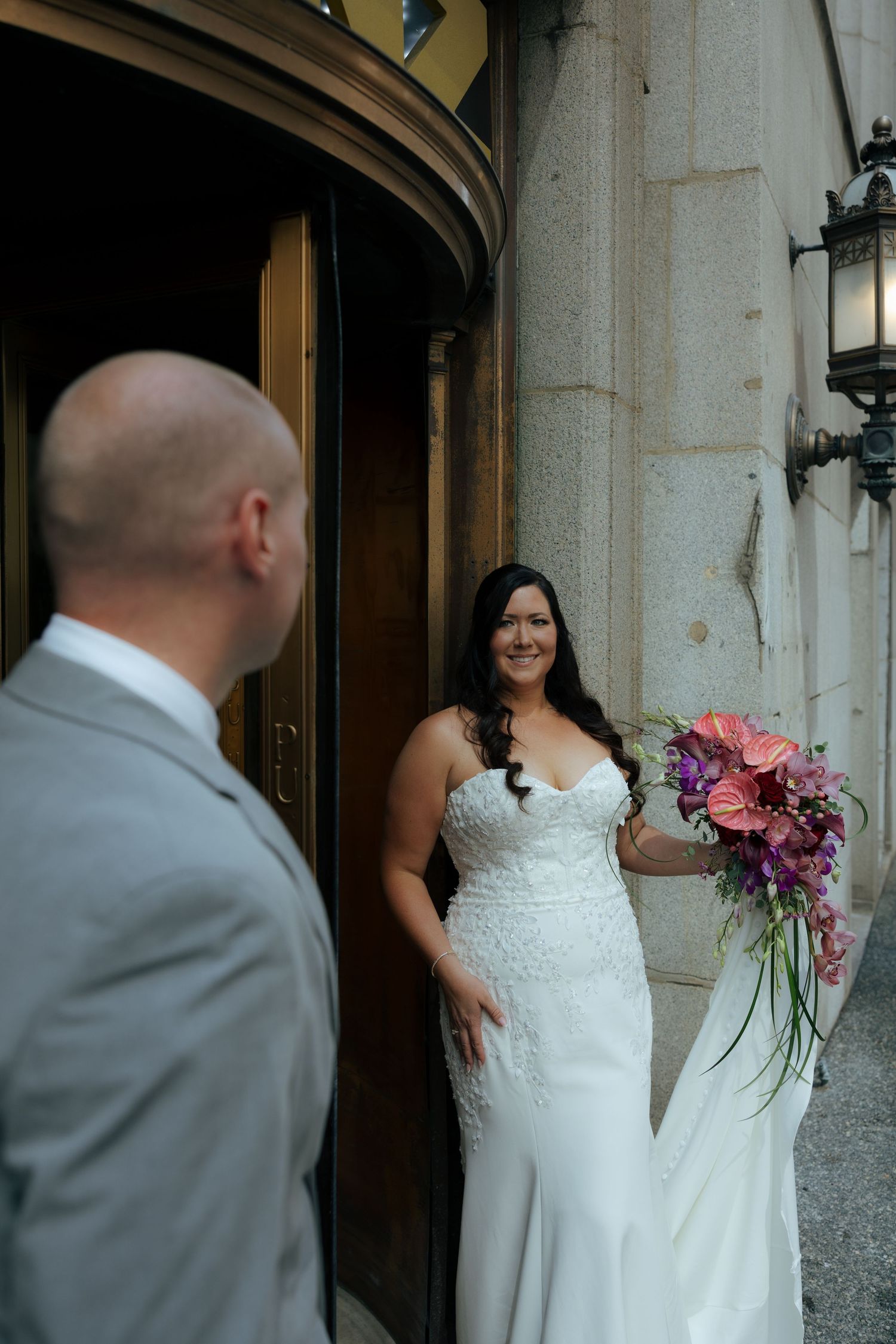 Bride in white strapless gown holding pink bouquet smiles at groom in gray suit outside elegant stone building.