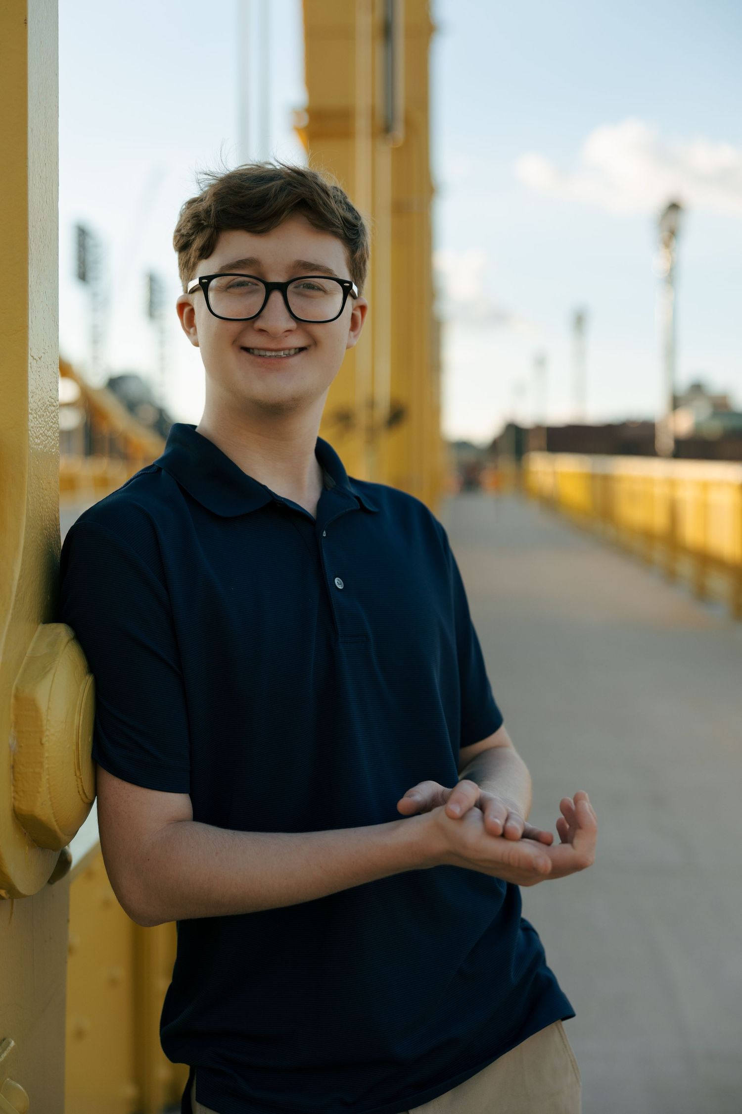 Smiling teenage boy with glasses in navy polo shirt leaning against yellow bridge railing outdoors.
