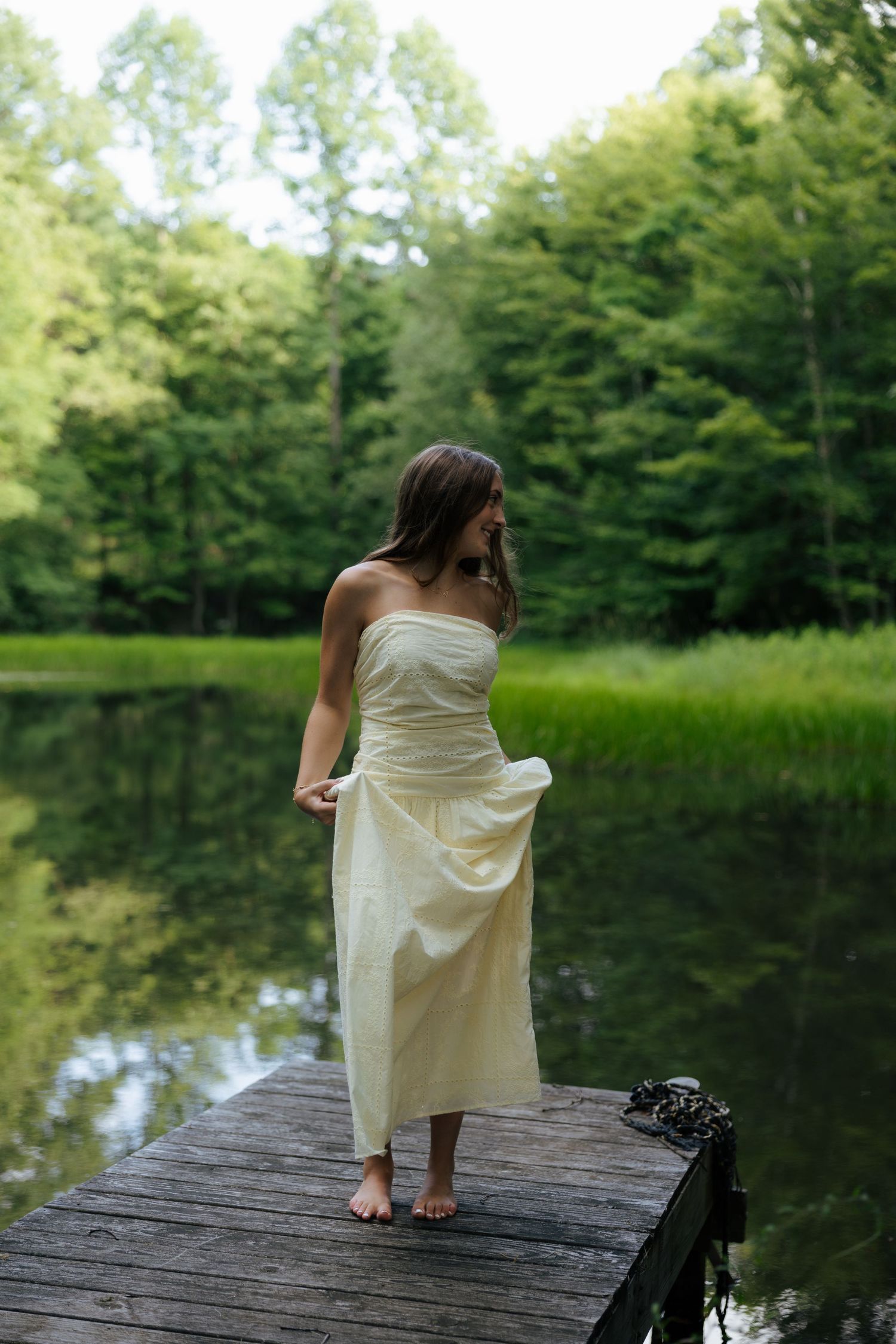 Young woman in white strapless dress standing barefoot on wooden dock by calm reflective lake.