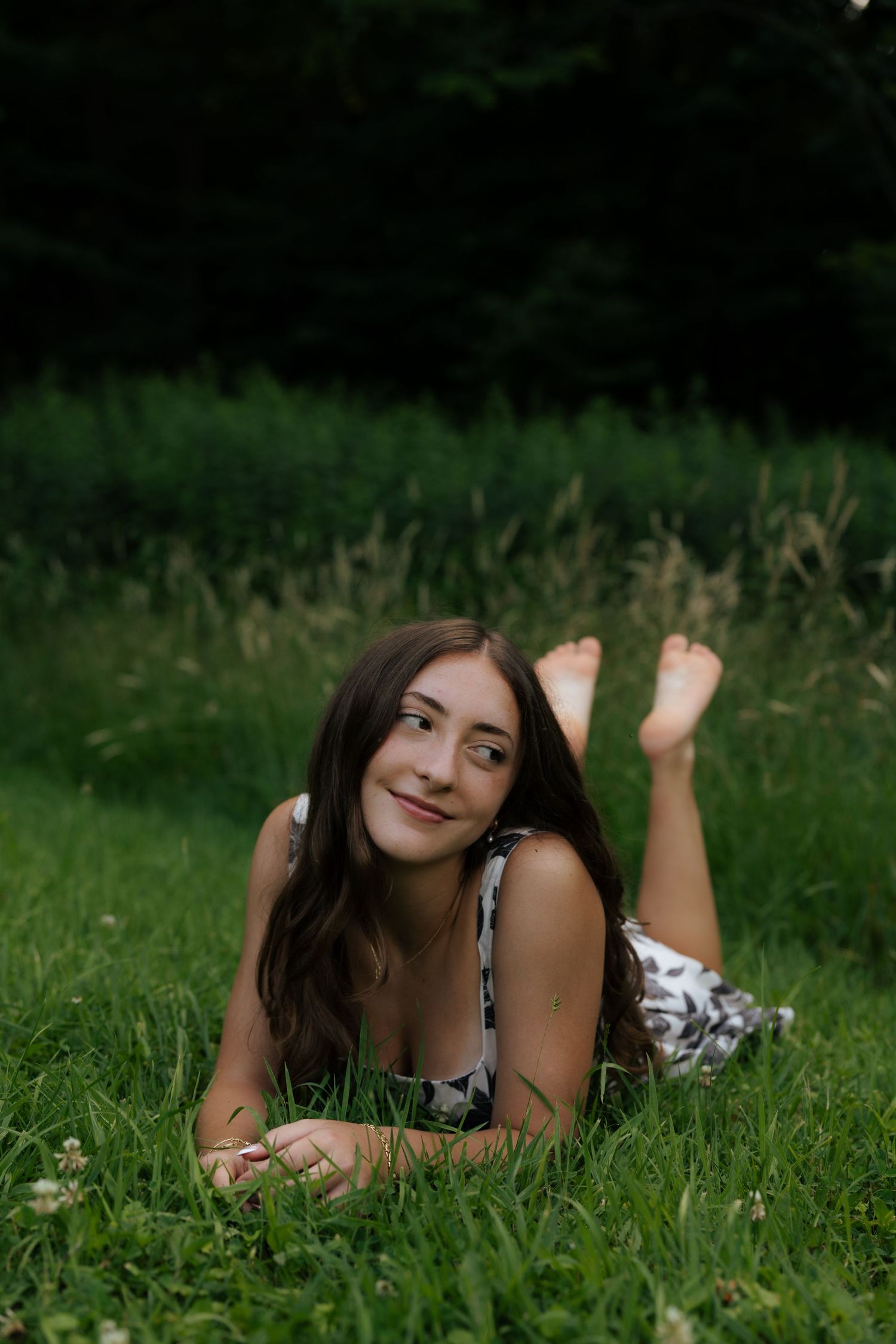 Young brunette woman lying barefoot on green grass smiling in a floral dress outdoors.