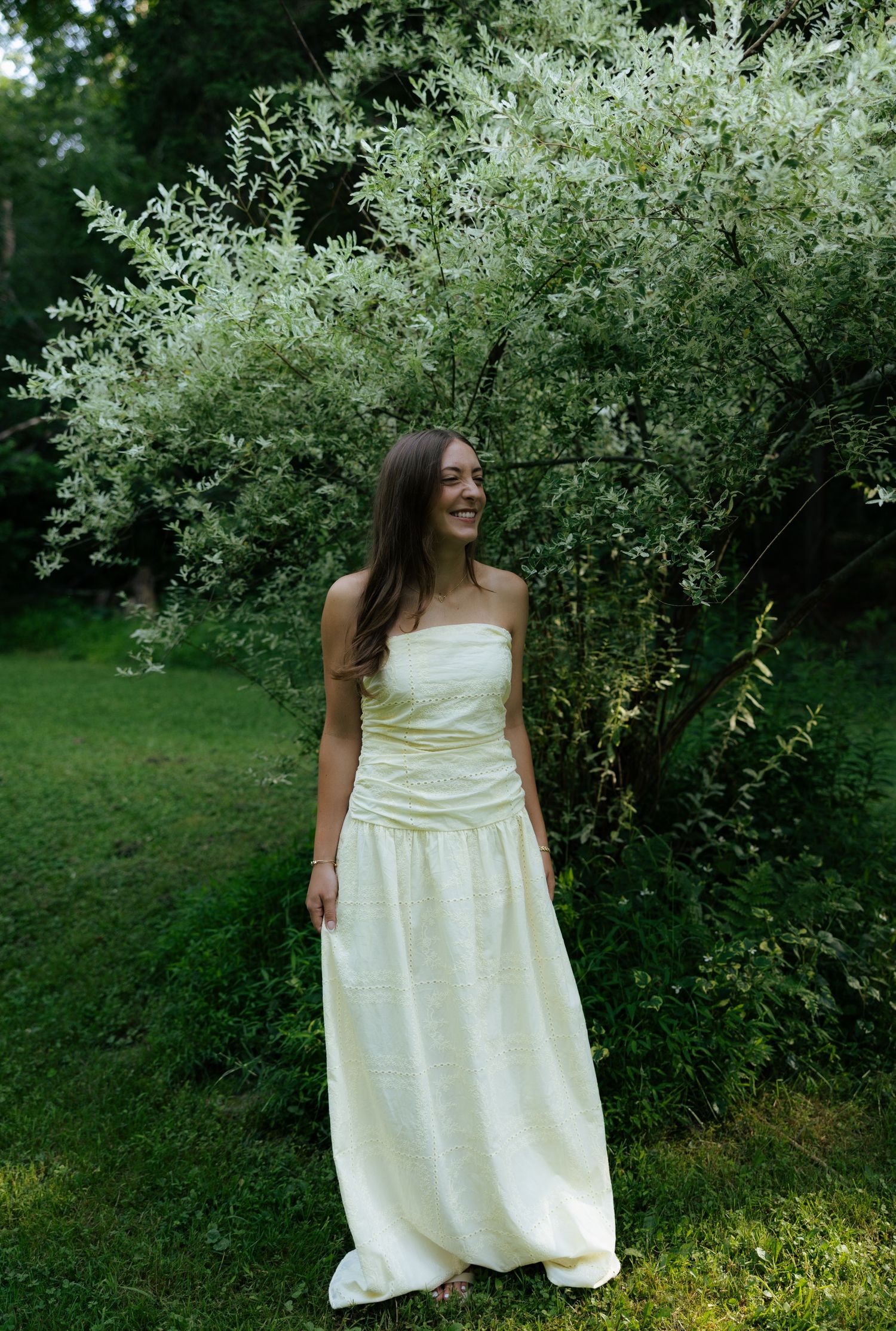 Young woman in cream strapless maxi dress standing before blooming white flowering shrub outdoors.