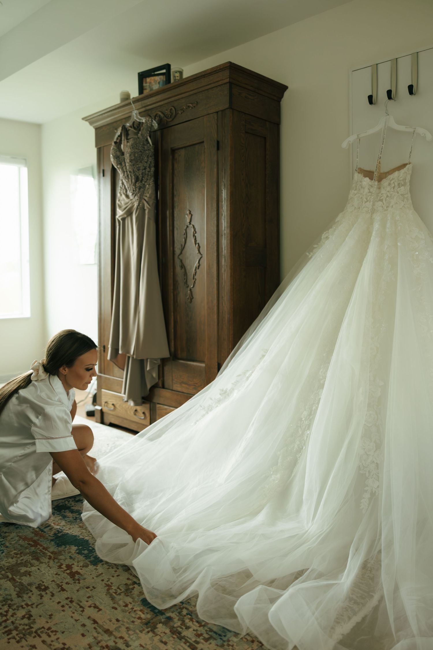 Bride arranging her elegant white lace wedding gown train in a rustic room with wooden armoire.