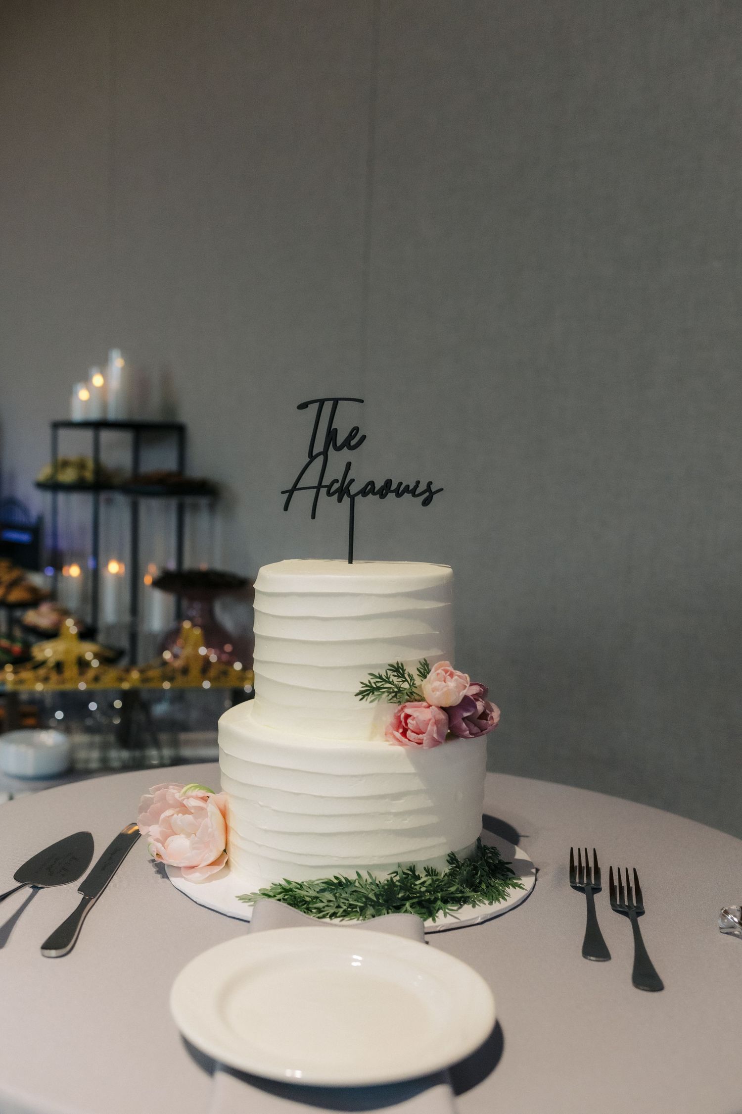Two-tier white textured wedding cake with pink flowers and The Achaows cake topper on a white table.