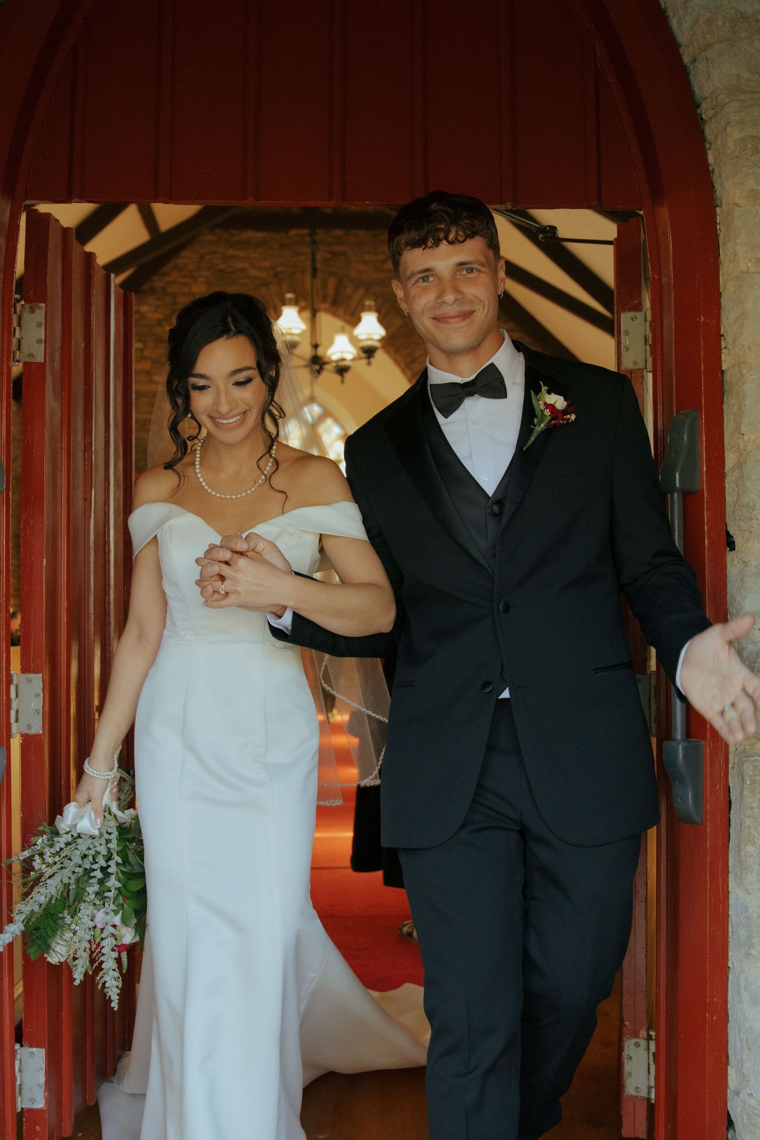 Newlyweds exit stone chapel through red arched doors, bride in white gown, groom in black tuxedo.