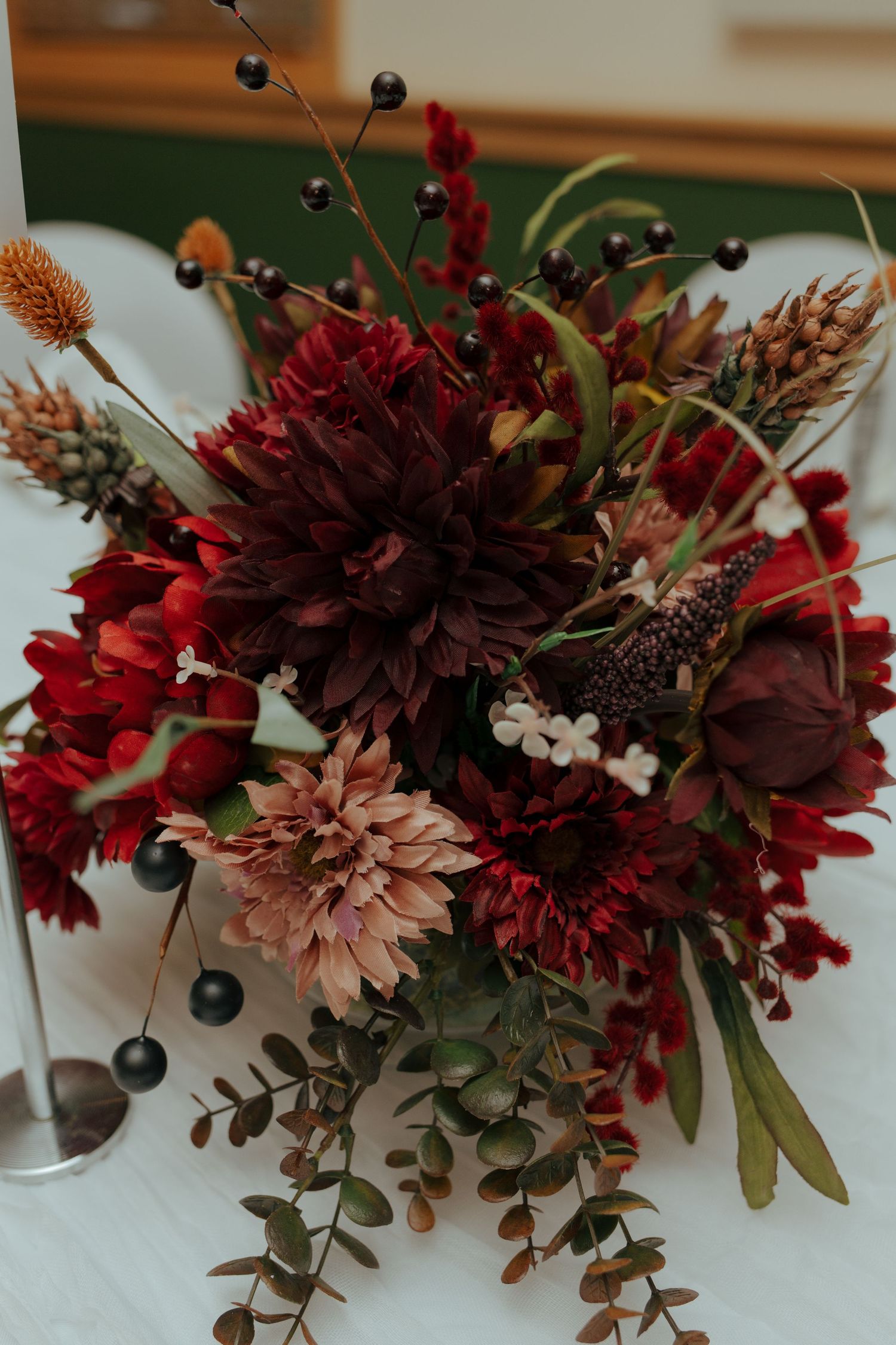 Stunning fall wedding bouquet featuring deep burgundy dahlias, red flowers, and dark berries on white table.