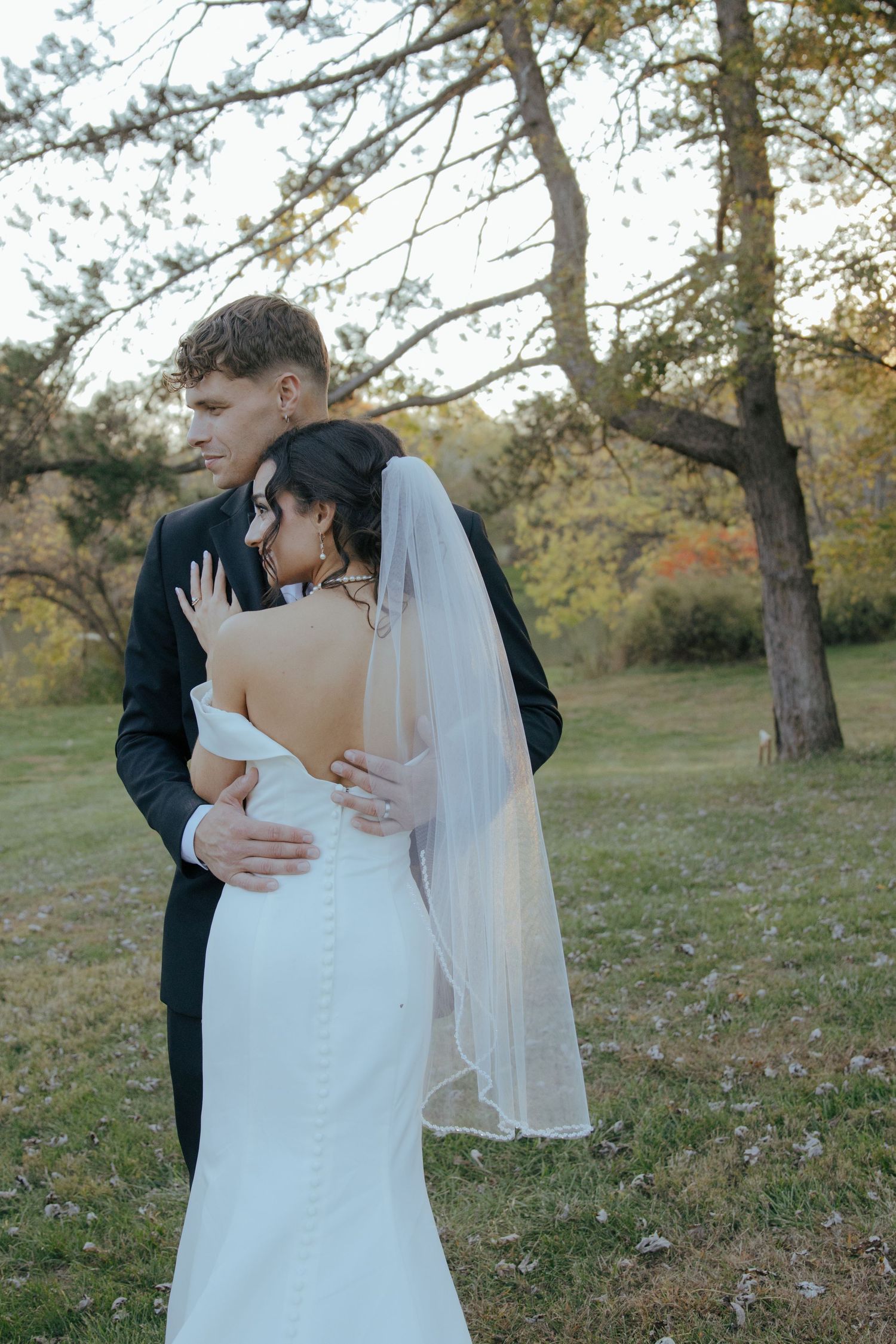 Groom embracing bride from behind in an autumn park, bride wearing backless mermaid gown and veil.