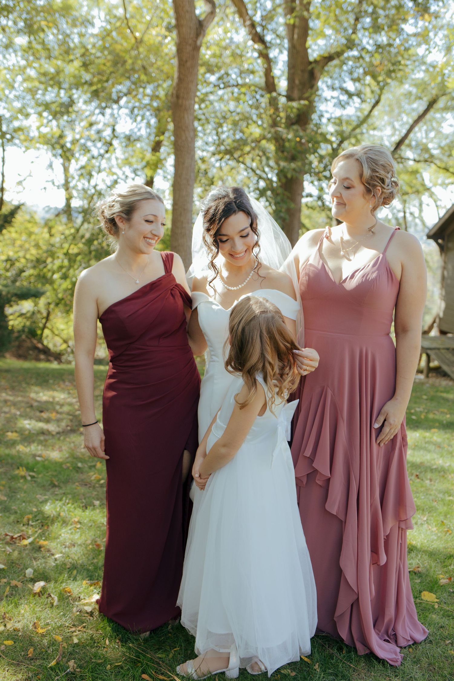 Bride in off-shoulder gown stands with two bridesmaids in burgundy and mauve dresses and a flower girl outdoors.