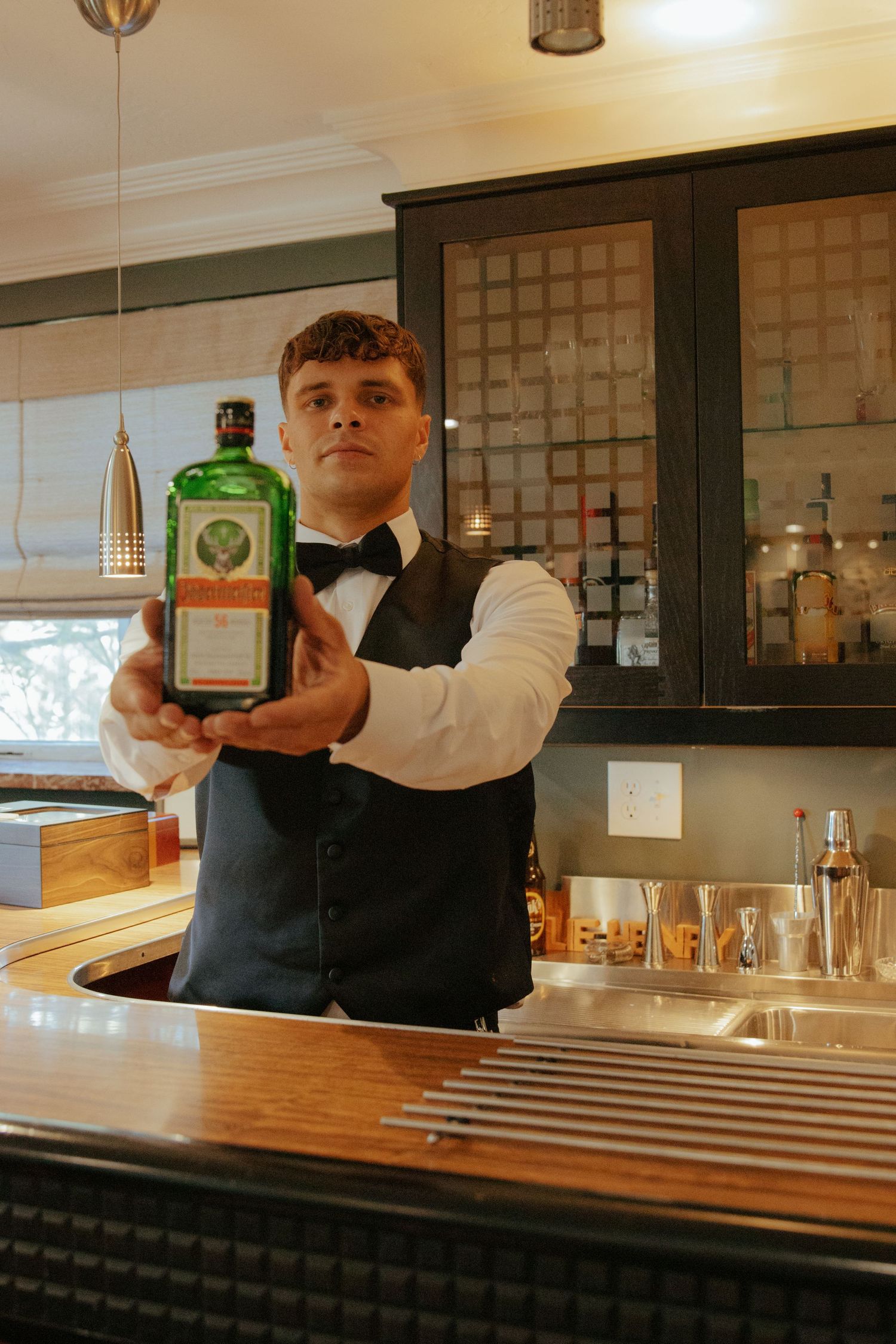 Bartender in vest and bow tie holds Jagermeister bottle toward camera at wedding reception bar.