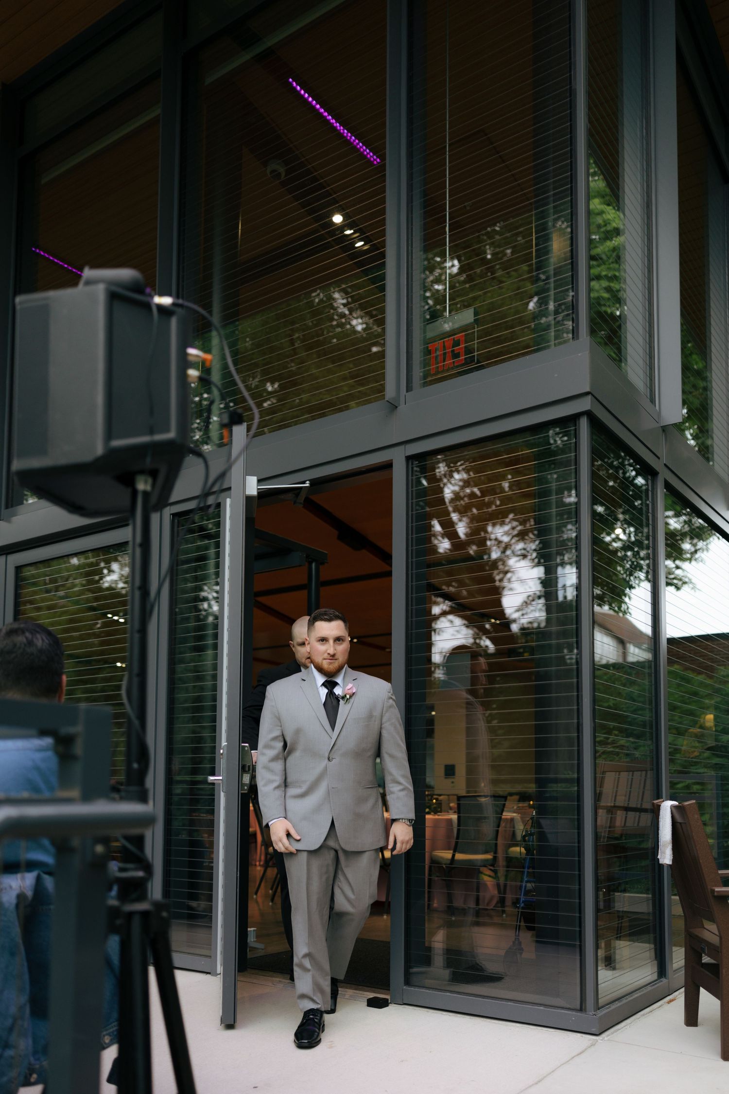 Groom in gray suit with boutonniere walks out of modern glass building doors at outdoor wedding venue.