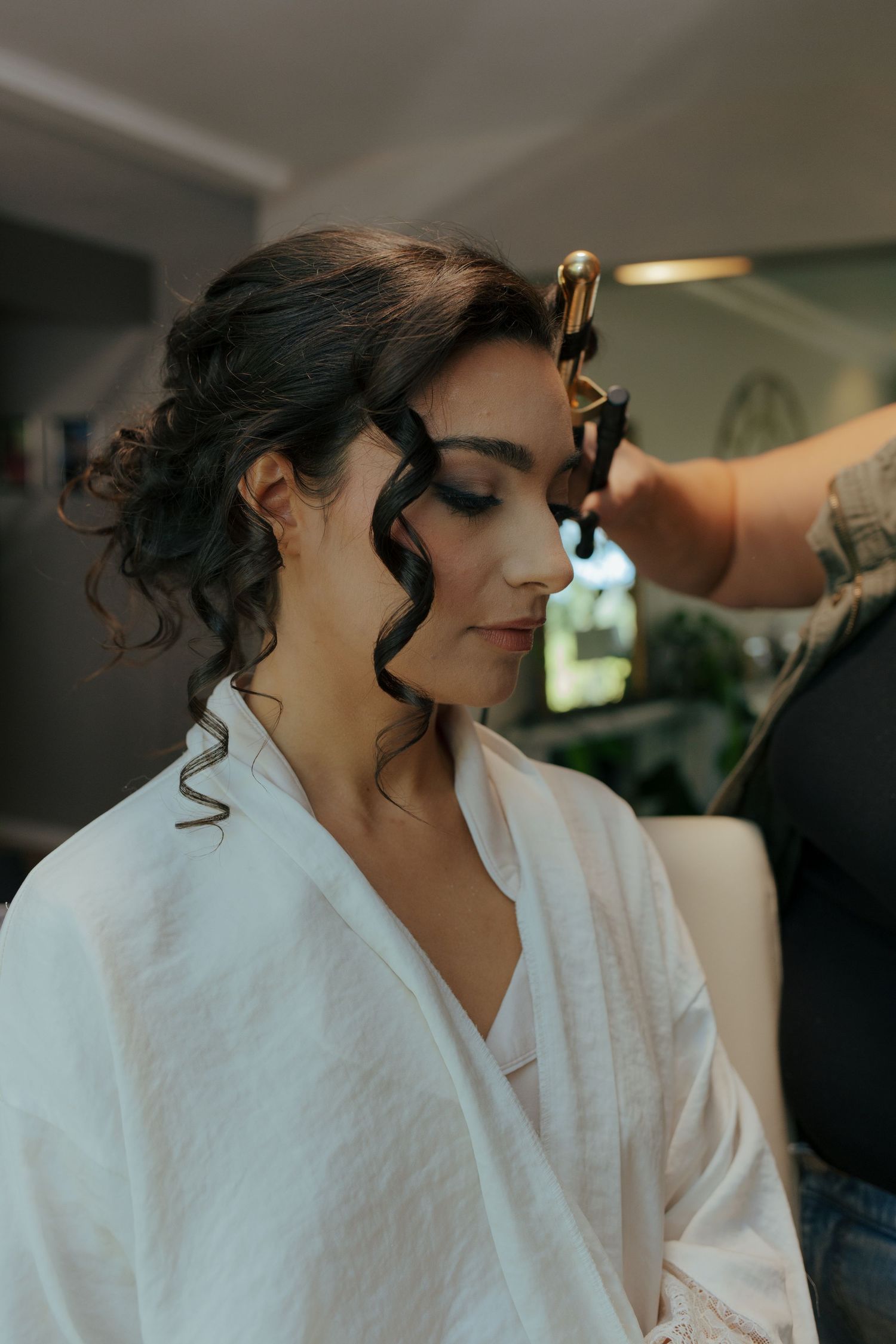 Bride in white robe getting curly updo hairstyle done by a stylist before her wedding ceremony.