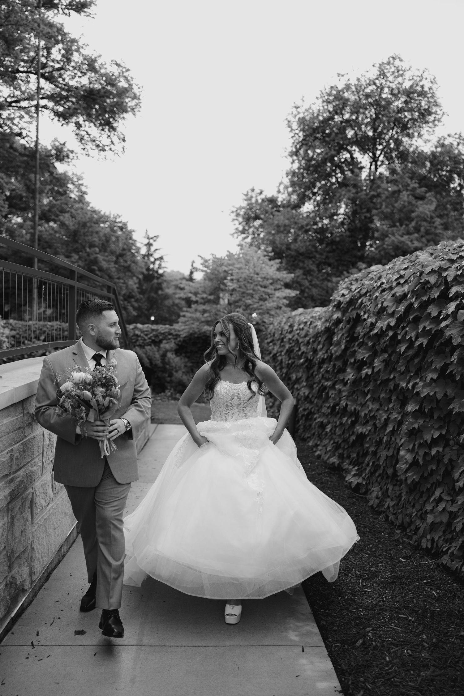 Black and white photo of a bride and groom walking together outdoors, surrounded by lush greenery on their wedding day.