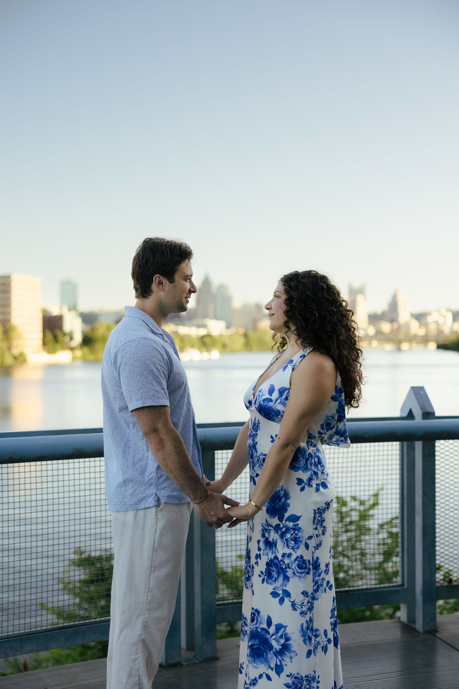 Couple holding hands facing each other on waterfront bridge with city skyline at sunset.