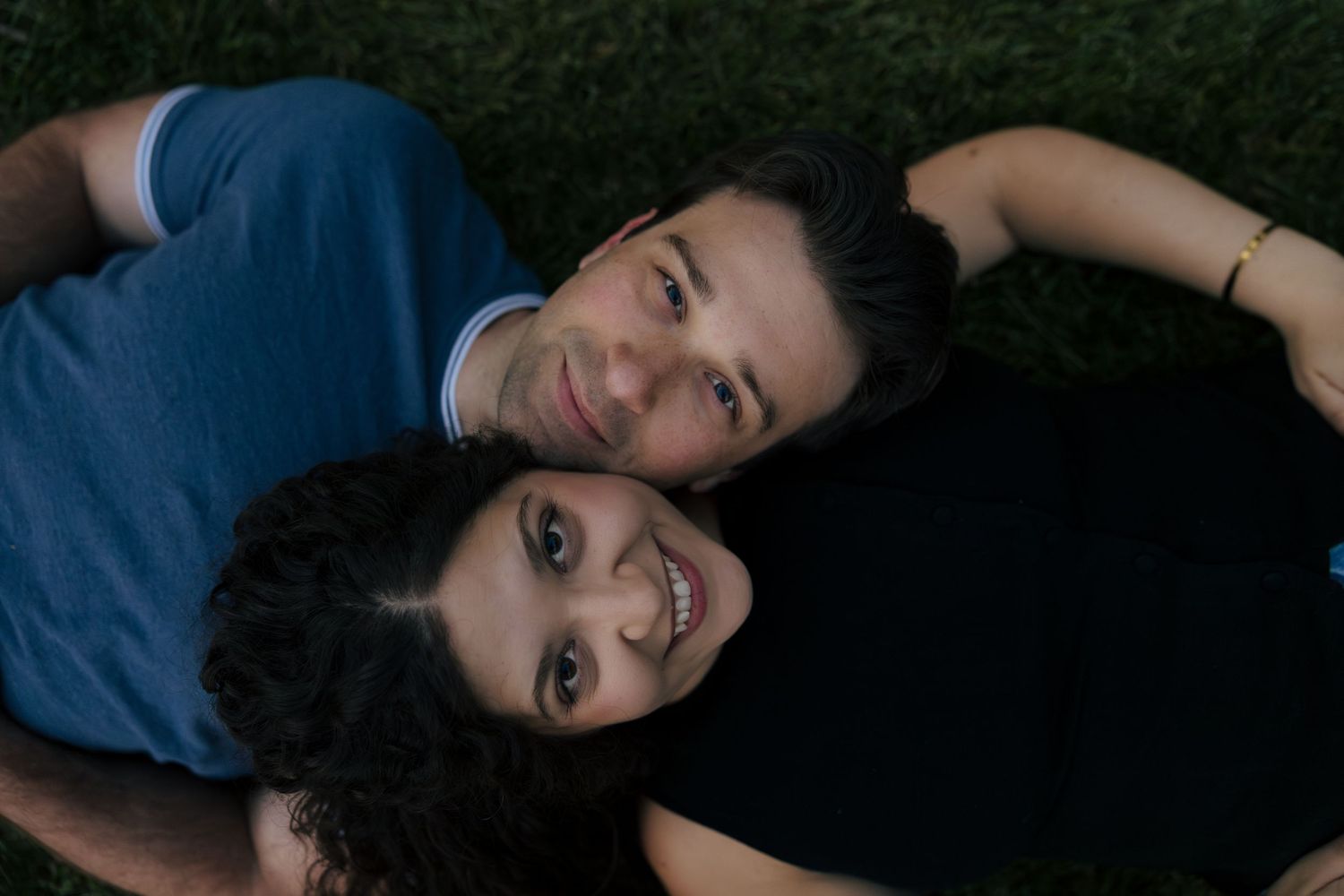 A smiling couple lies on green grass, looking up at the camera in a relaxed and happy outdoor portrait.