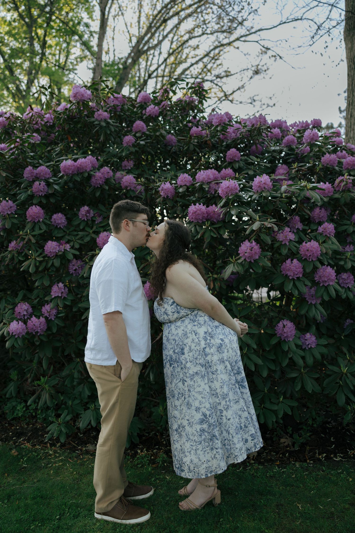 Couple kissing in front of large purple rhododendron bush in a garden park.