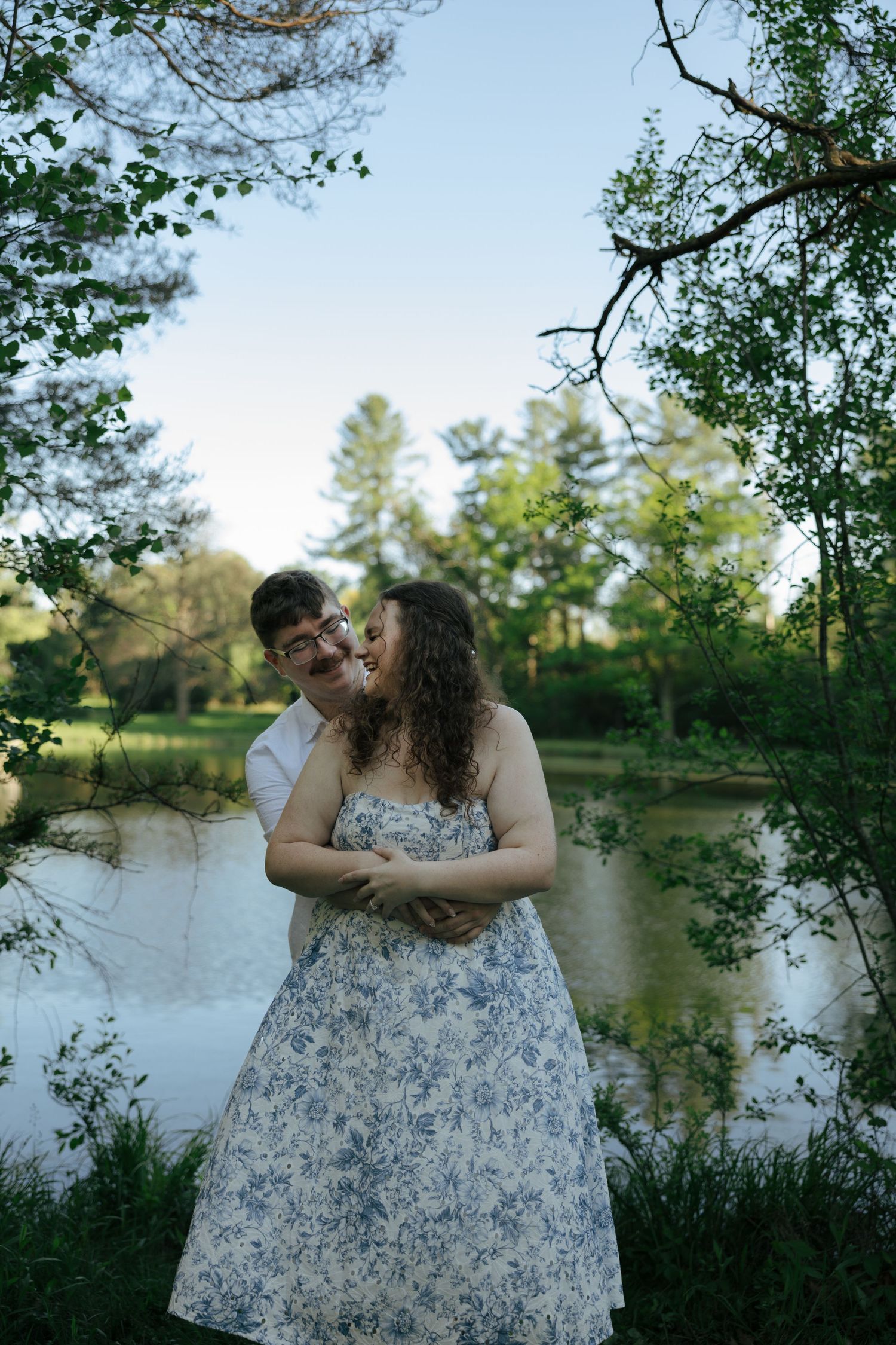 Engaged couple laughing and embracing near a reflective pond, framed by green trees and branches at dusk.