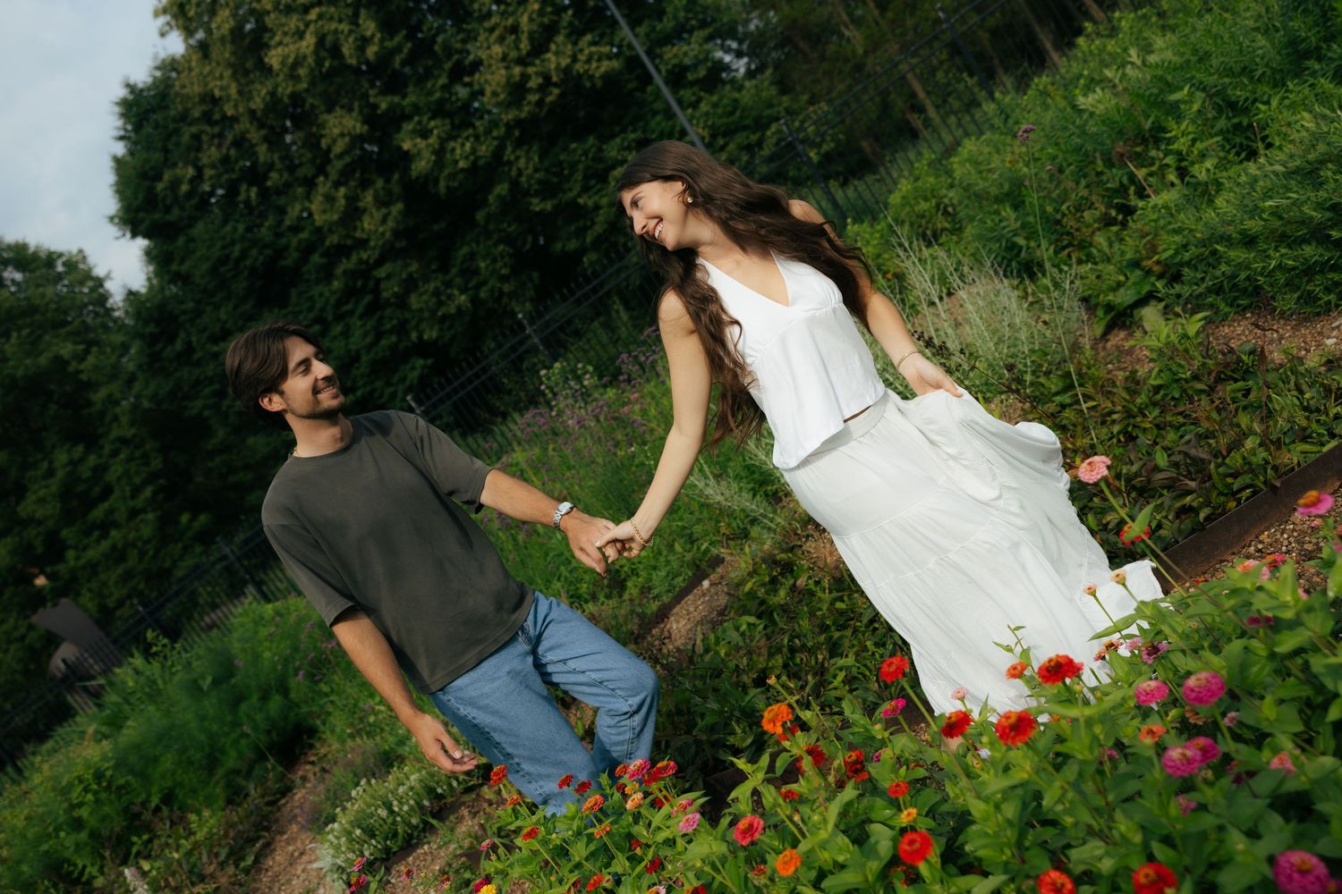 A joyful couple holds hands and walks through a vibrant garden filled with colorful flowers on a summer day.