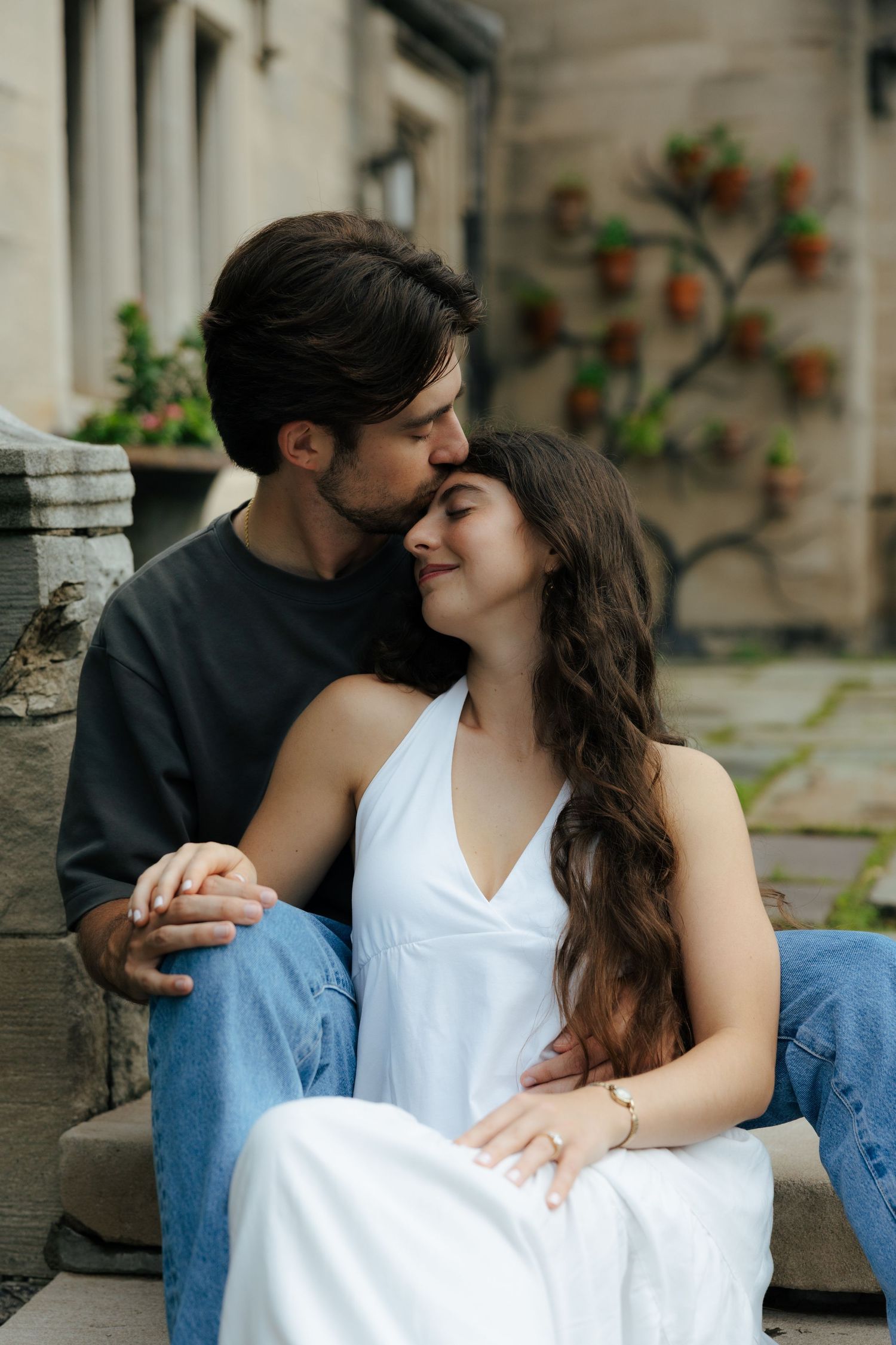 Couple sitting on stone steps sharing a tender kiss with potted plants in the background.