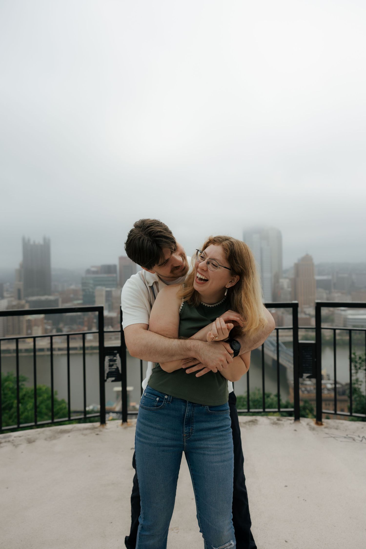 Happy couple hugging and laughing on an outdoor overlook with a foggy urban skyline in the background.