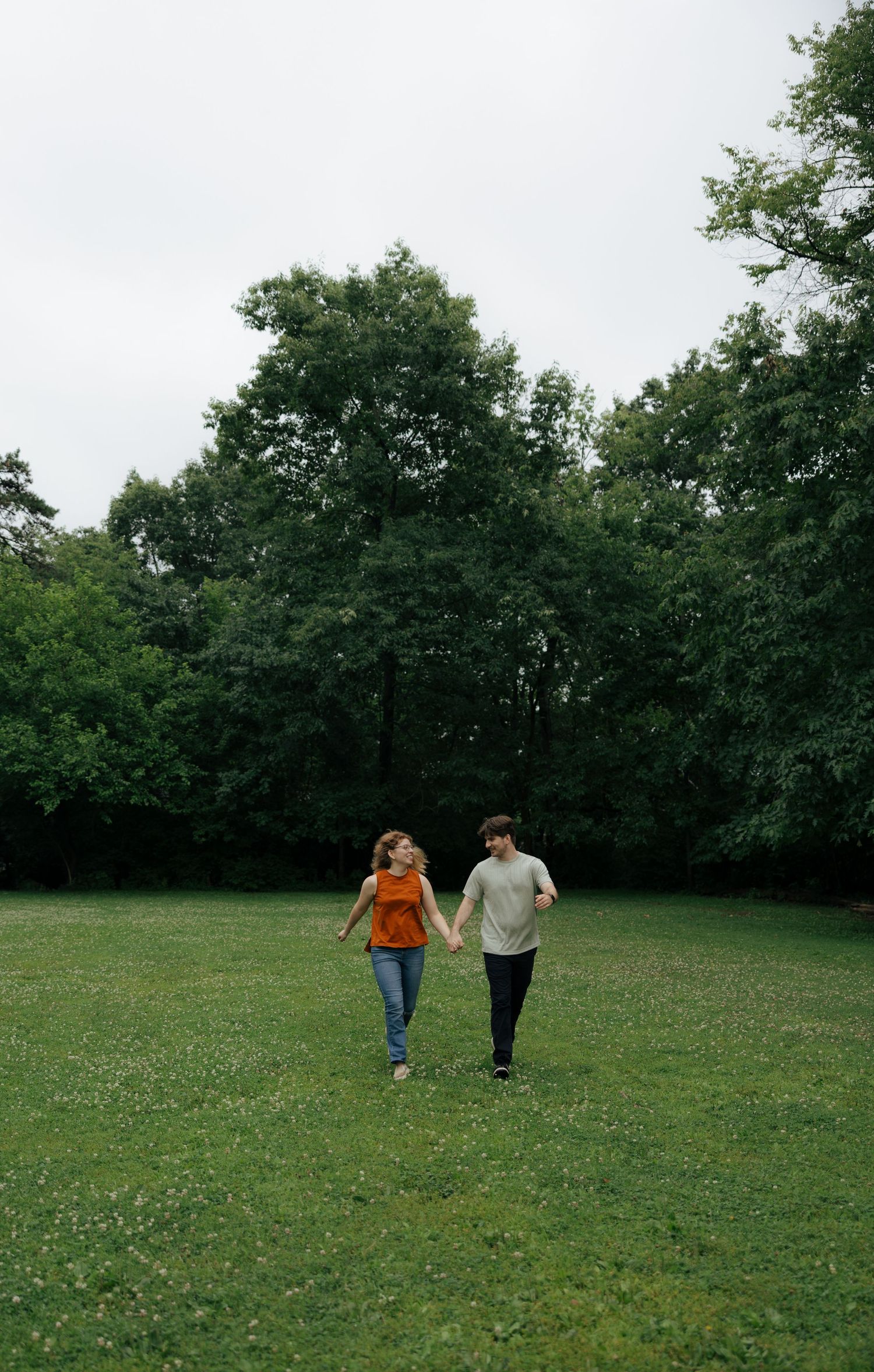 A couple holds hands while walking through a wide open green park under an overcast sky.
