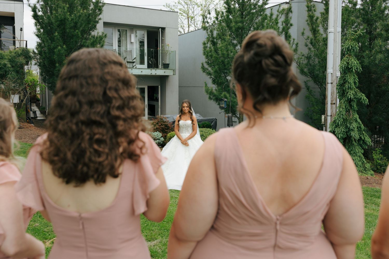 Bridesmaids in pink dresses watch a bride in a white gown walk toward them outdoors near a modern building.