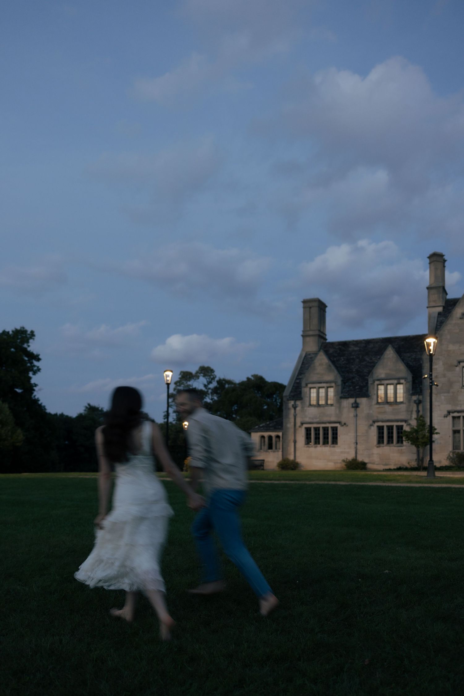 Couple holding hands running across a green lawn at twilight with an elegant stone estate in background.