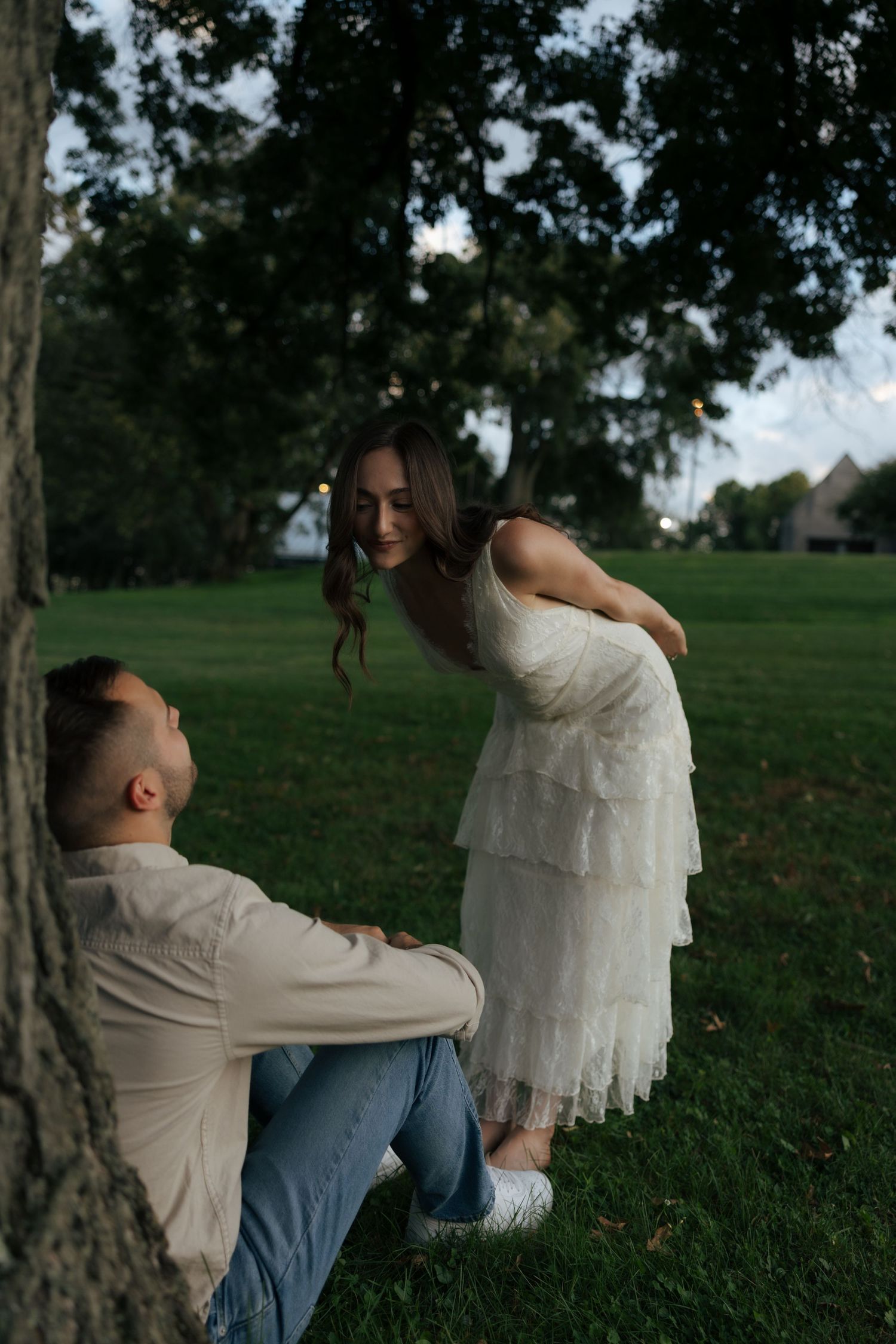 A woman in a white tiered dress leans toward a man sitting against a large tree in a lush green park.