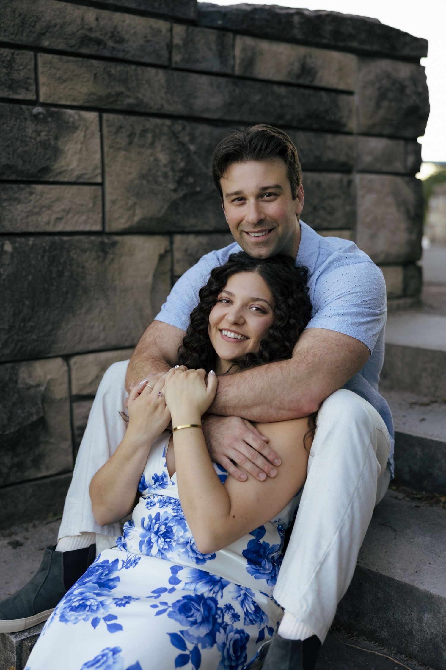 A happy couple poses on stone steps, with the man embracing the woman from behind, both smiling warmly.