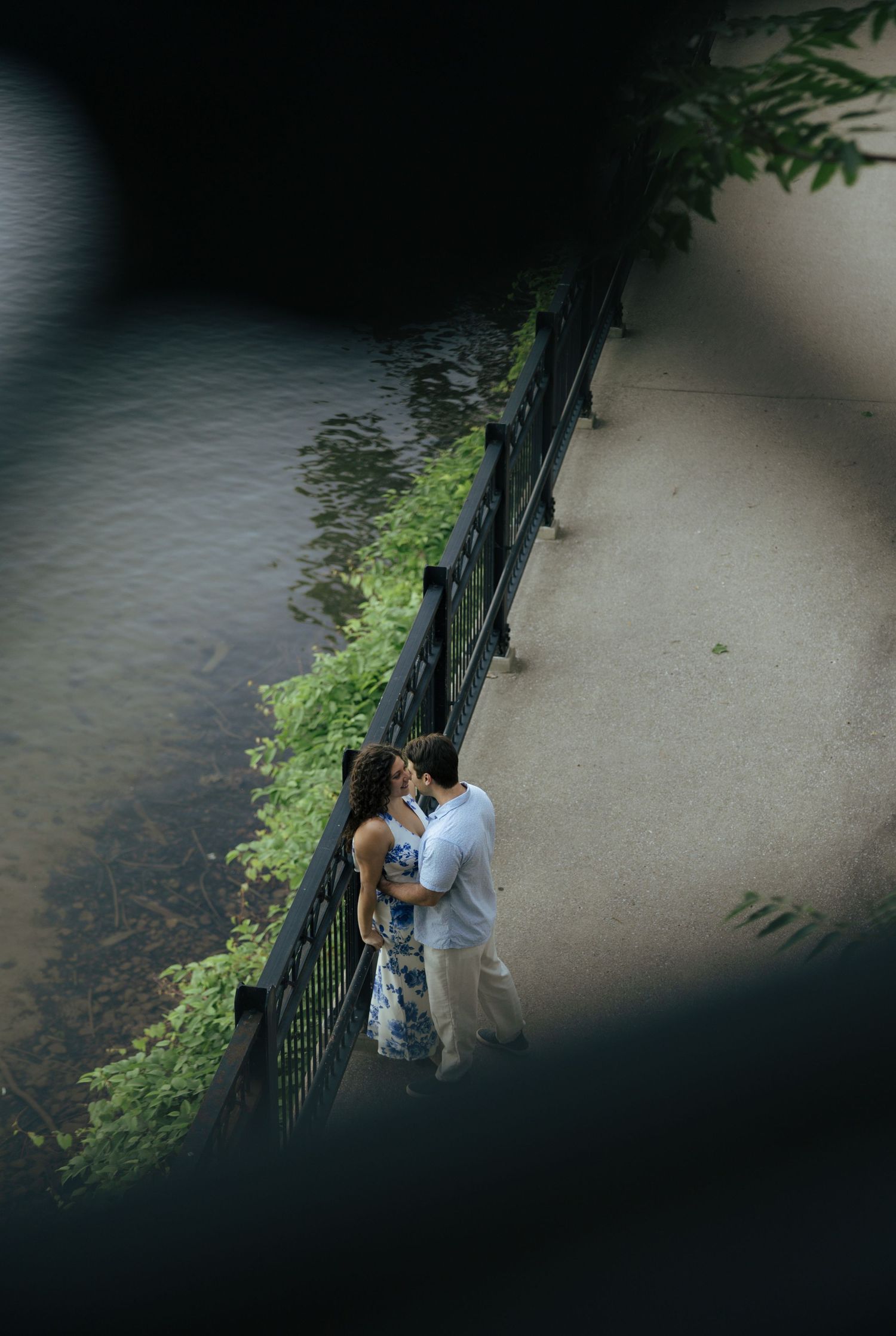An aerial view of a couple sharing a romantic moment along a riverside walkway with green water beside them.