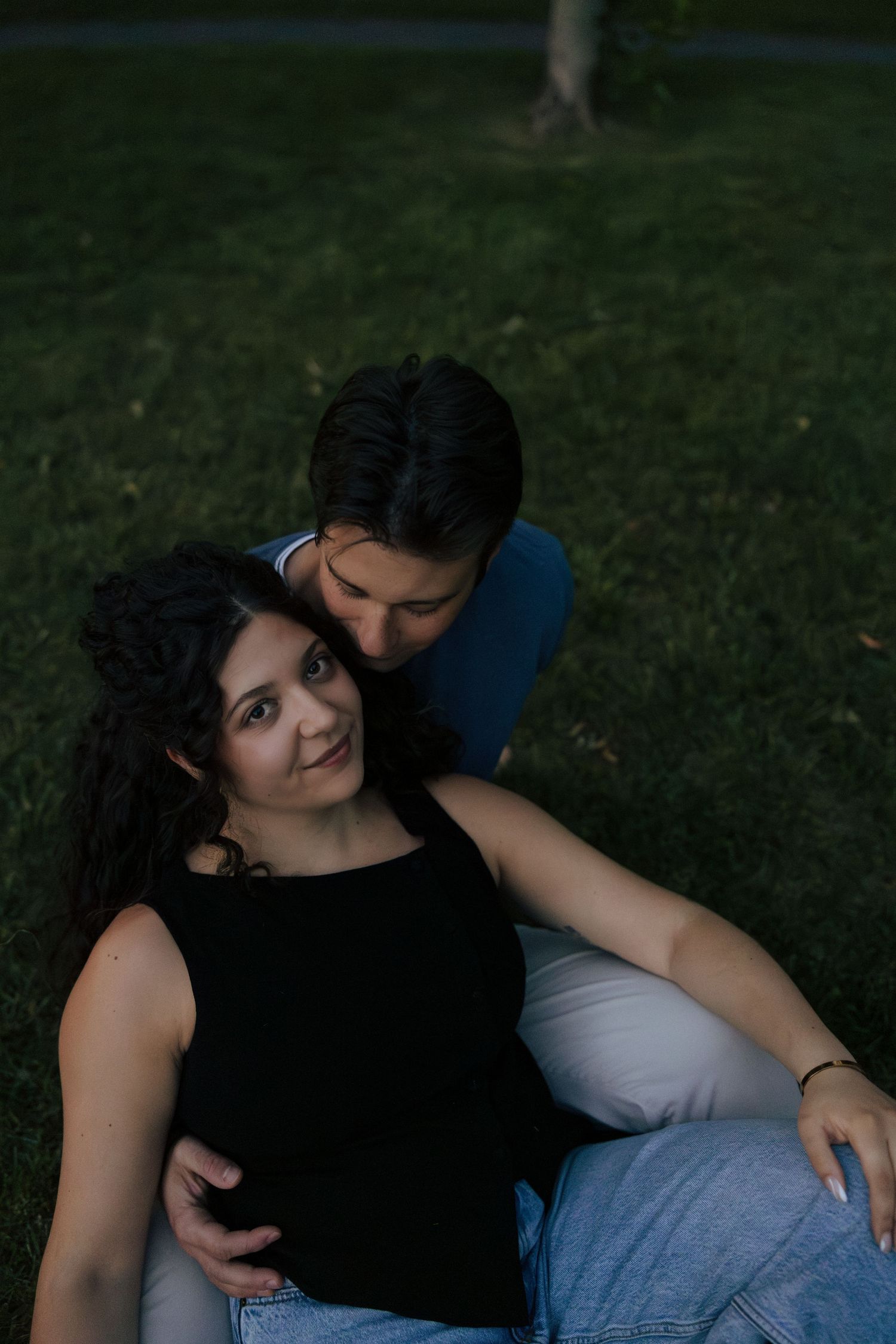 Couple relaxing on grass at dusk, woman leaning back against man who kisses her head.