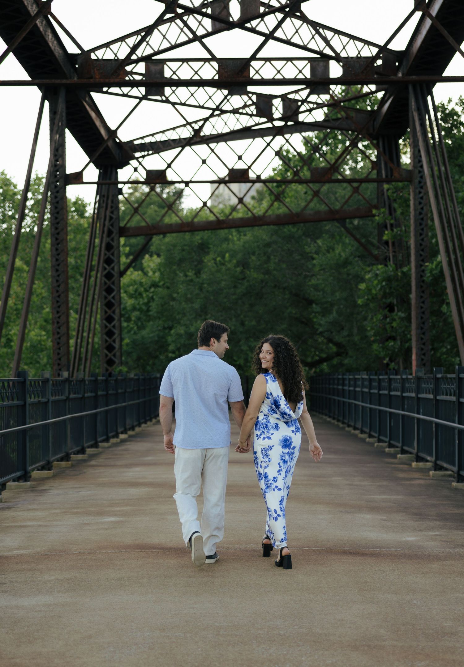 An engaged couple walks hand-in-hand through a historic iron truss bridge surrounded by lush green trees.